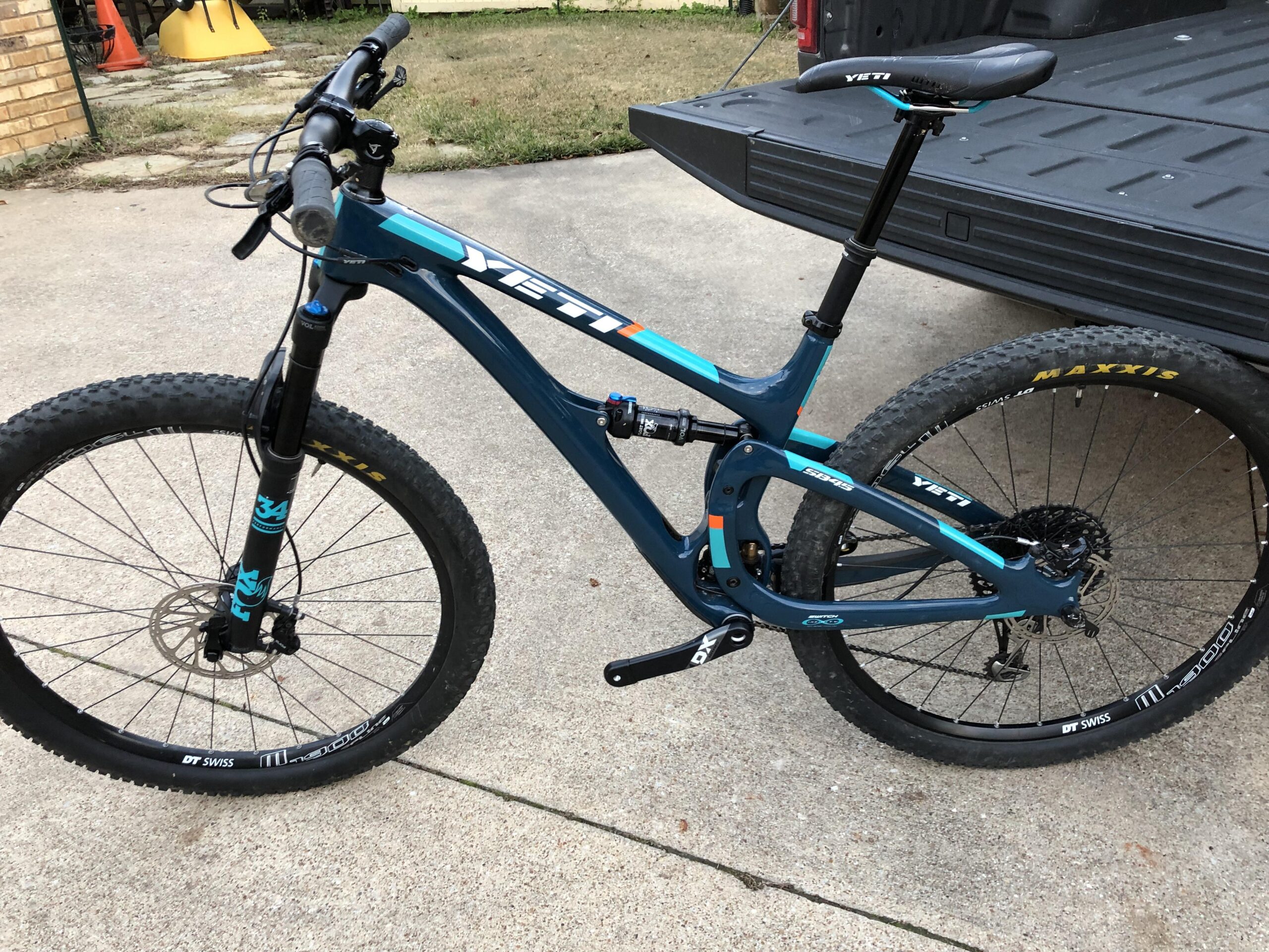 Yeti SB4.5c: A blue Yeti mountain bike is parked beside a pickup truck. The bike features thick tires designed for off-road riding, a sleek frame with vibrant accents, and a dropper seat post. The background shows a concrete surface and some grass.