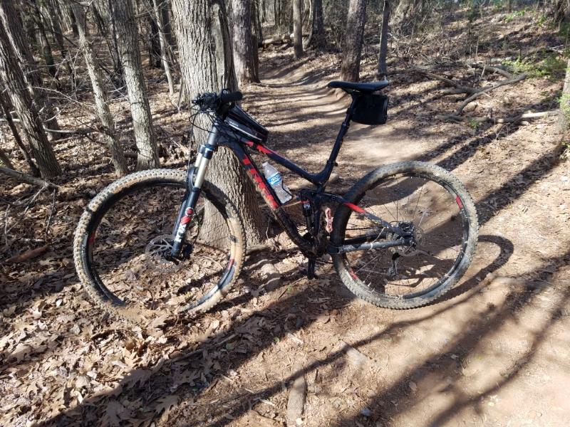 Trek Fuel EX 8: A black and red mountain bike parked on a dirt path surrounded by trees and fallen leaves in a wooded area. Parts of the bike are muddy, indicating recent use on trails. Sunlight filters through the trees, casting shadows on the ground.