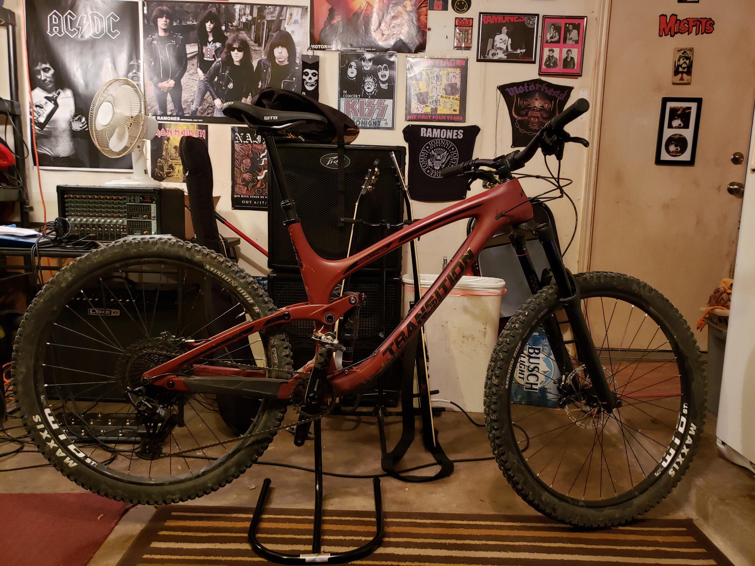 Transition Sentinel: A mountain bike is displayed in a cluttered room adorned with various rock band posters. The bike is positioned on a stand, showcasing its vibrant red frame and thick tires. In the background, there are amplifiers, a fan, and a doorway with decorative elements, adding to the eclectic vibe of the space.