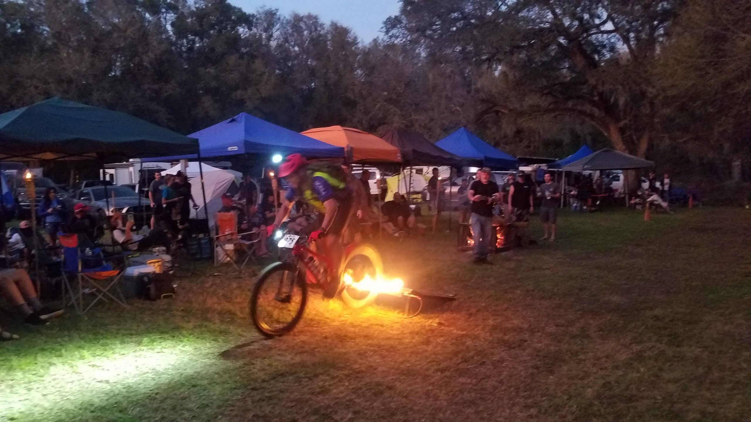 A mountain biker is performing a jump over flames during an event, surrounded by tents and spectators in a grassy area at twilight. The scene captures the excitement of the activity and the ambiance created by the warm light of the fire against the evening sky. Imba Red mountain bike trail.