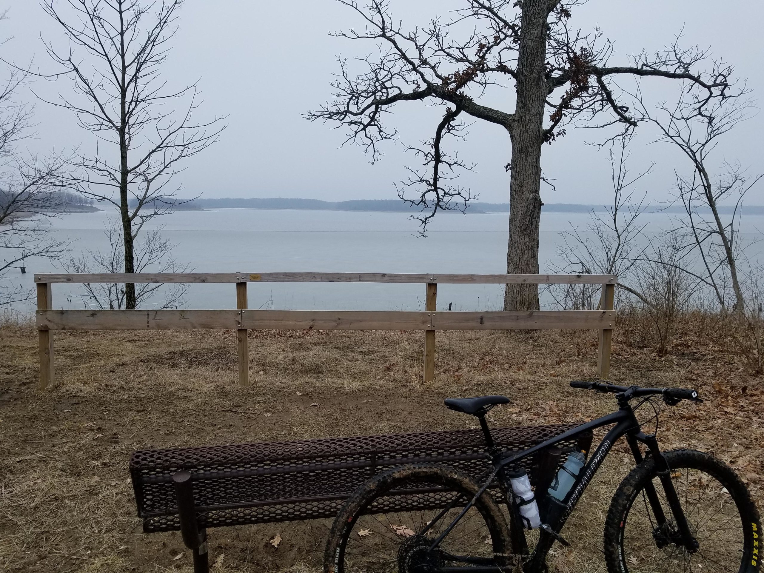 A scenic view of a calm lake on a cloudy day, with a wooden fence and two bare trees in the foreground. A brown bench is positioned near the water's edge, and a mountain bike rests alongside it, partially covered in mud. The surrounding landscape features dry grass and a misty atmosphere. Camp Camfield mountain bike trail.