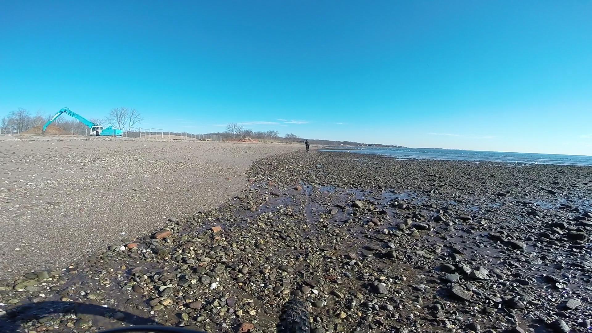 A rocky shoreline with a person walking along the beach under a clear blue sky. In the background, a construction vehicle is seen working near a fenced area. The water gently laps at the shore, reflecting the bright daylight. Wolfes Pond park mountain bike trail.