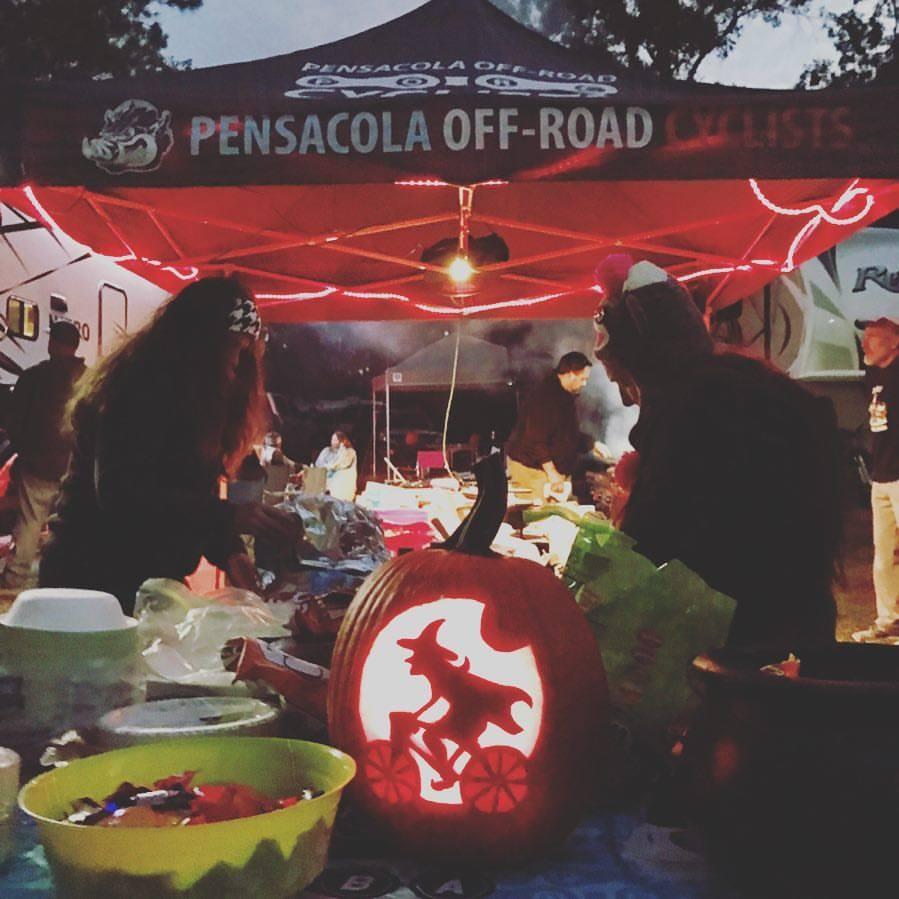 A festive outdoor gathering featuring a cooking area under a tent labeled "Pensacola Off-Road Cyclists." In the foreground, a carved pumpkin depicting a witch riding a bicycle is illuminated. People are engaged in various activities, with tables set up with food and decorations, creating a lively atmosphere in the evening light. UWF Mountain Bike Trails mountain bike trail.