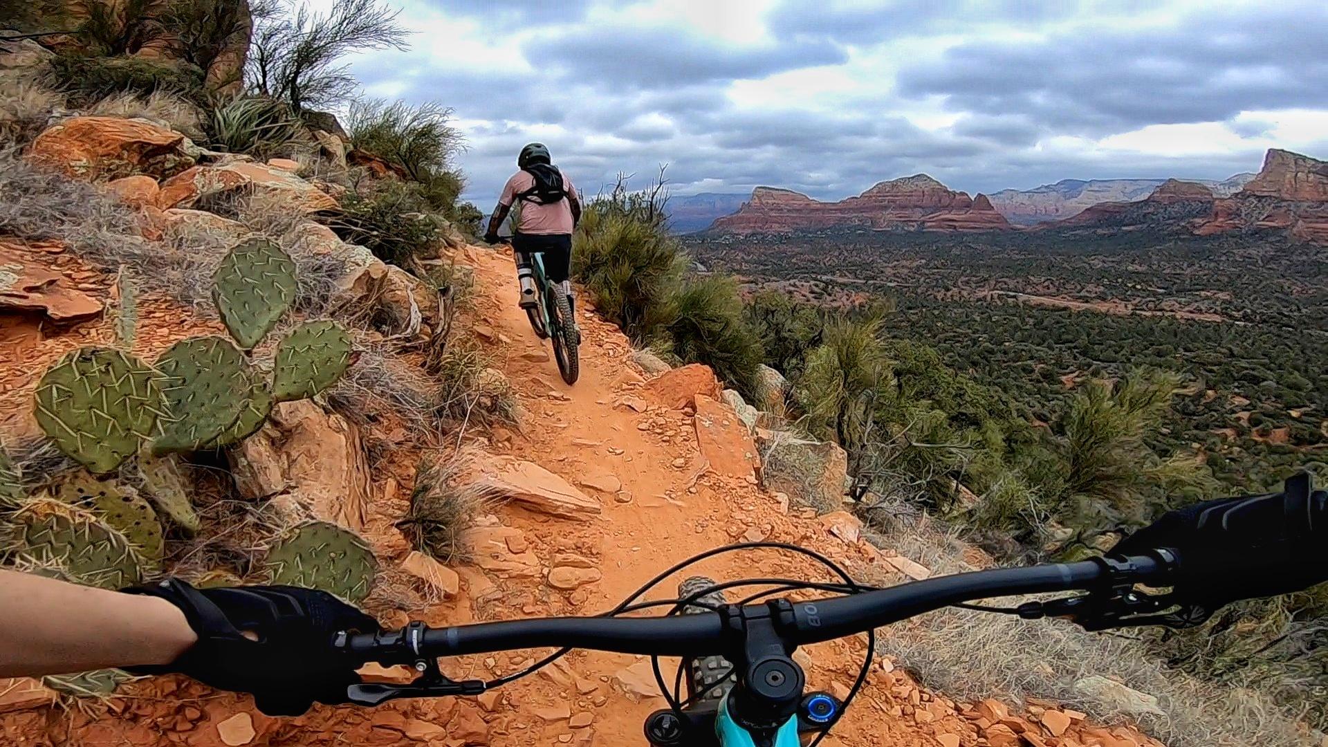A mountain biker navigates a rocky trail surrounded by desert vegetation, including cacti. The scene features dramatic red rock formations and a vast landscape under a cloudy sky. The perspective captures the biker from the front, highlighting the handlebars of the bike and the rugged terrain. Hiline mountain bike trail.