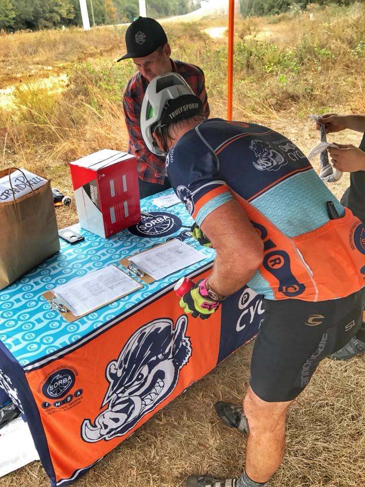 A cyclist wearing a brightly colored jersey and helmet is leaning over a table with clipboards and a red box. Another person, wearing a plaid shirt and cap, is watching nearby. The table has a banner featuring an illustrated wild boar logo, and the background shows a grassy area with trees. UWF Mountain Bike Trails mountain bike trail.