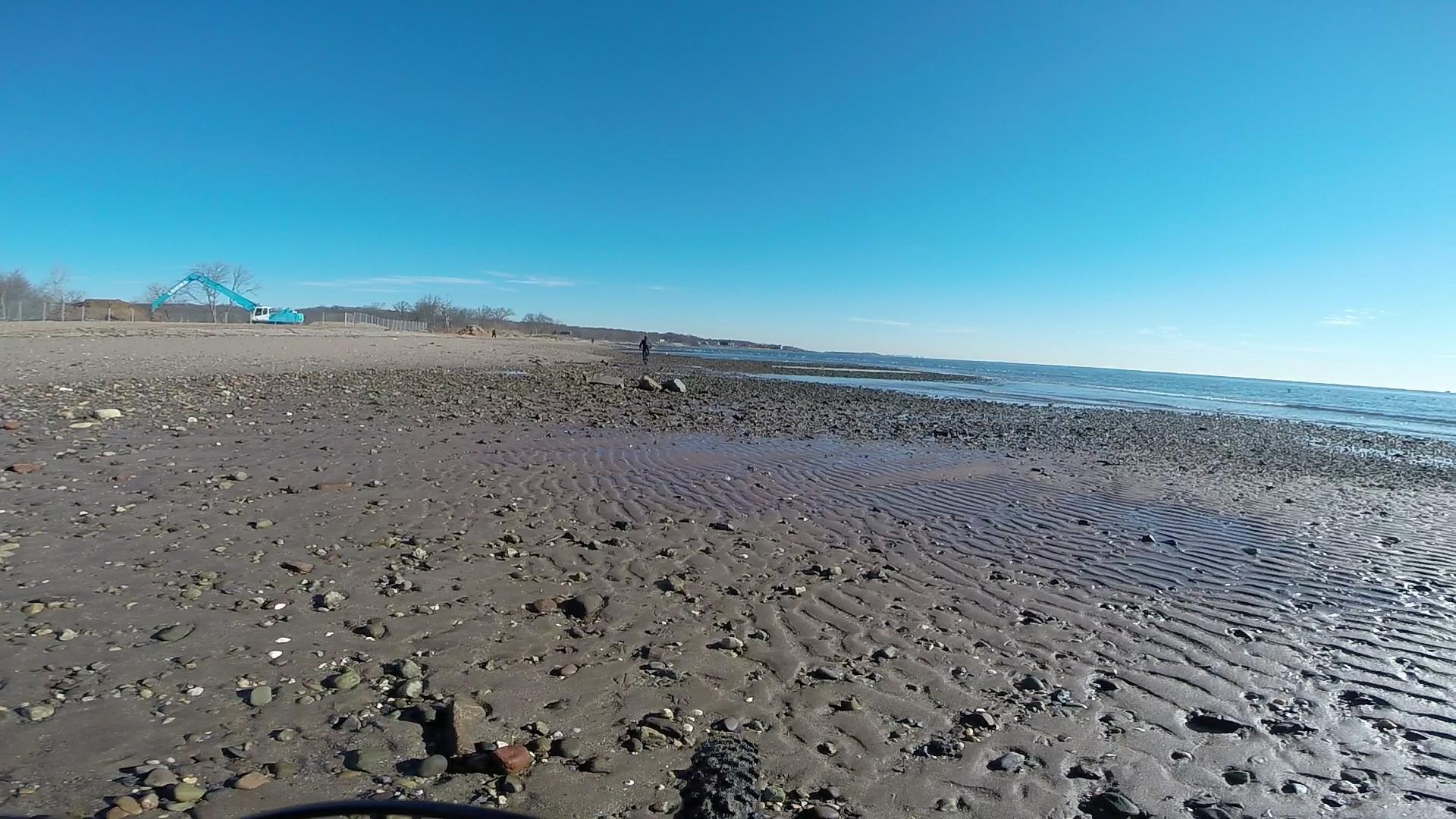 A sandy beach with scattered pebbles and rocks, under a clear blue sky. In the background, a person walks along the shoreline, and a light blue construction vehicle is visible near a fenced area. The water reflects the sun, creating a serene coastal scene. Wolfes Pond park mountain bike trail.