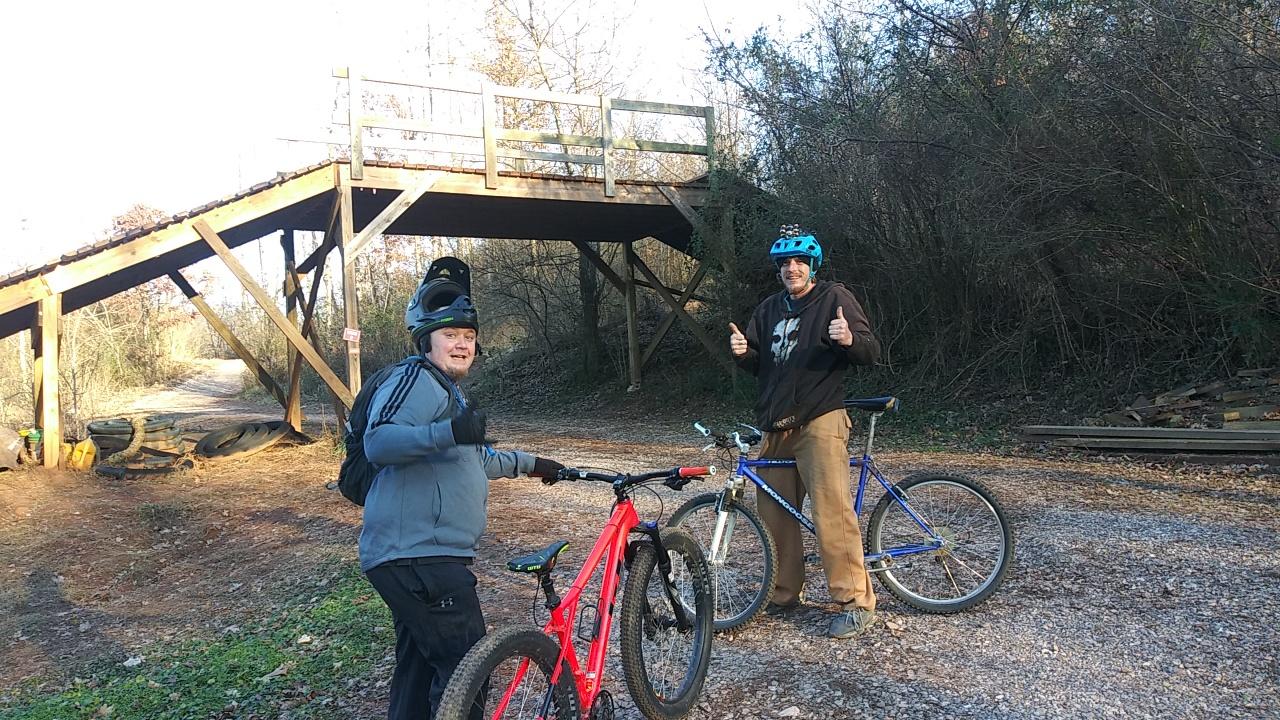 GT Ricochet: Two mountain bikers stand with their bikes on a gravel path near a wooden bridge in a wooded area. One rider is wearing a light gray jacket and helmet, giving a thumbs-up, while the other, in a brown hoodie, also smiles and shows a thumbs-up. The background features greenery and a sunny sky.