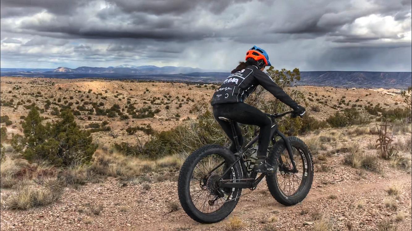 A cyclist riding a fat tire bike on a rugged trail in a desert landscape. The cyclist is wearing a black outfit and a blue helmet, and is facing away from the camera as they navigate a rocky path. In the background, dramatic gray clouds loom over distant mountains and arid terrain, highlighting the vastness of the outdoor setting. Mariposa Fat Bike Trails mountain bike trail.