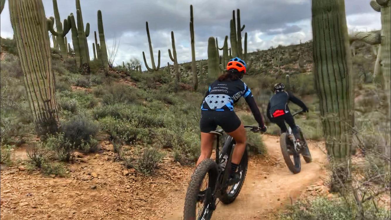 Two mountain bikers ride along a dirt trail surrounded by cacti and desert vegetation under a cloudy sky. One rider is in a blue and black outfit with an orange helmet, while the other is wearing a black outfit with a red accent. The terrain is rocky, with patches of greenery and sandy ground visible. Sweetwater Preserve mountain bike trail.