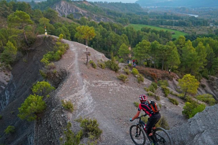 A mountain biker navigates a rocky path on a hillside, surrounded by lush green trees and distant mountains under a sunset sky. The trail is narrow and winding, leading toward a distant viewpoint where other outdoor enthusiasts can be seen.