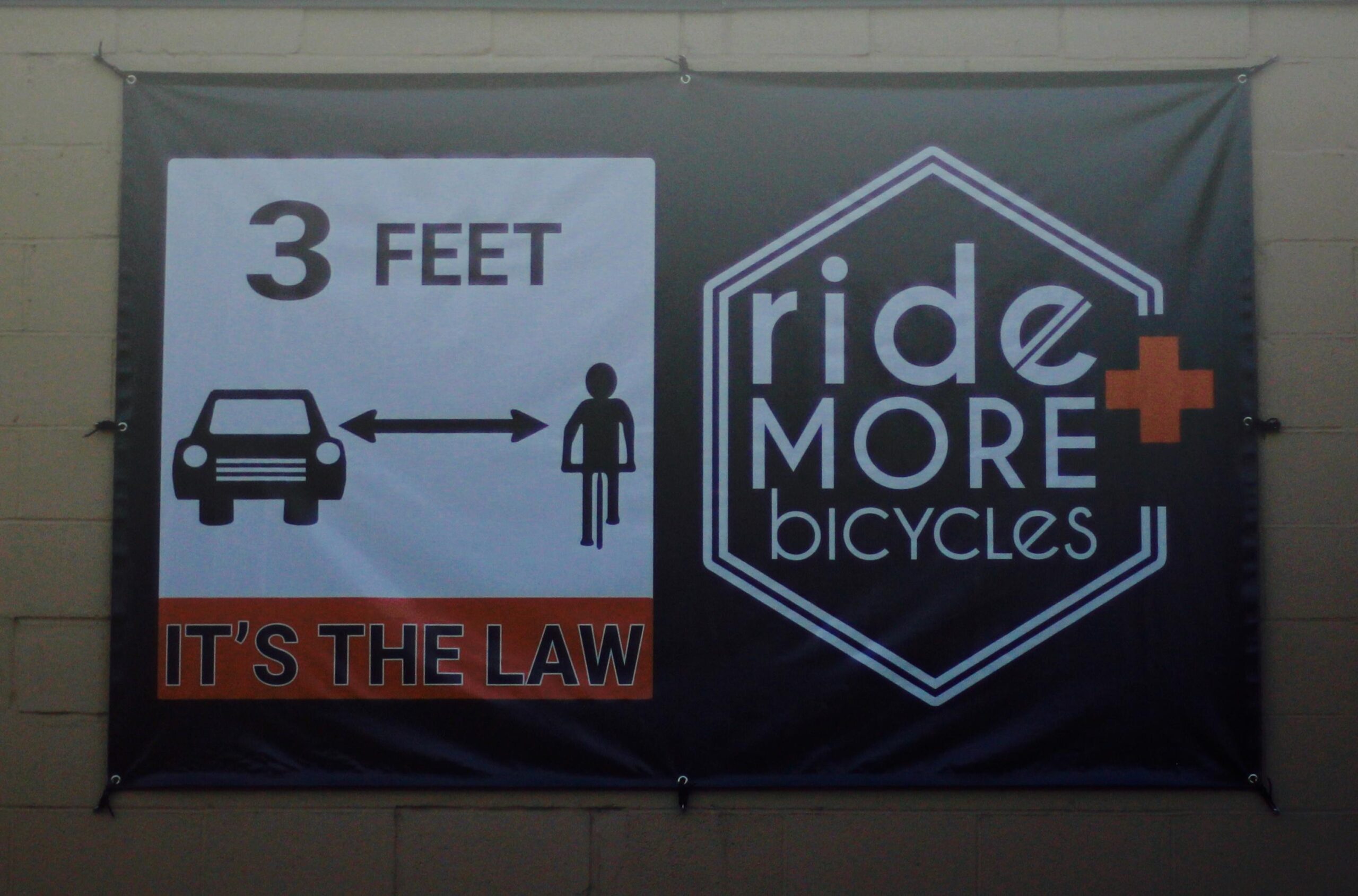 A banner displaying a graphic with a car and a person, emphasizing the requirement of maintaining a distance of 3 feet between vehicles and cyclists. The text reads "3 FEET IT'S THE LAW," alongside the logo for "ride MORE bicycles."