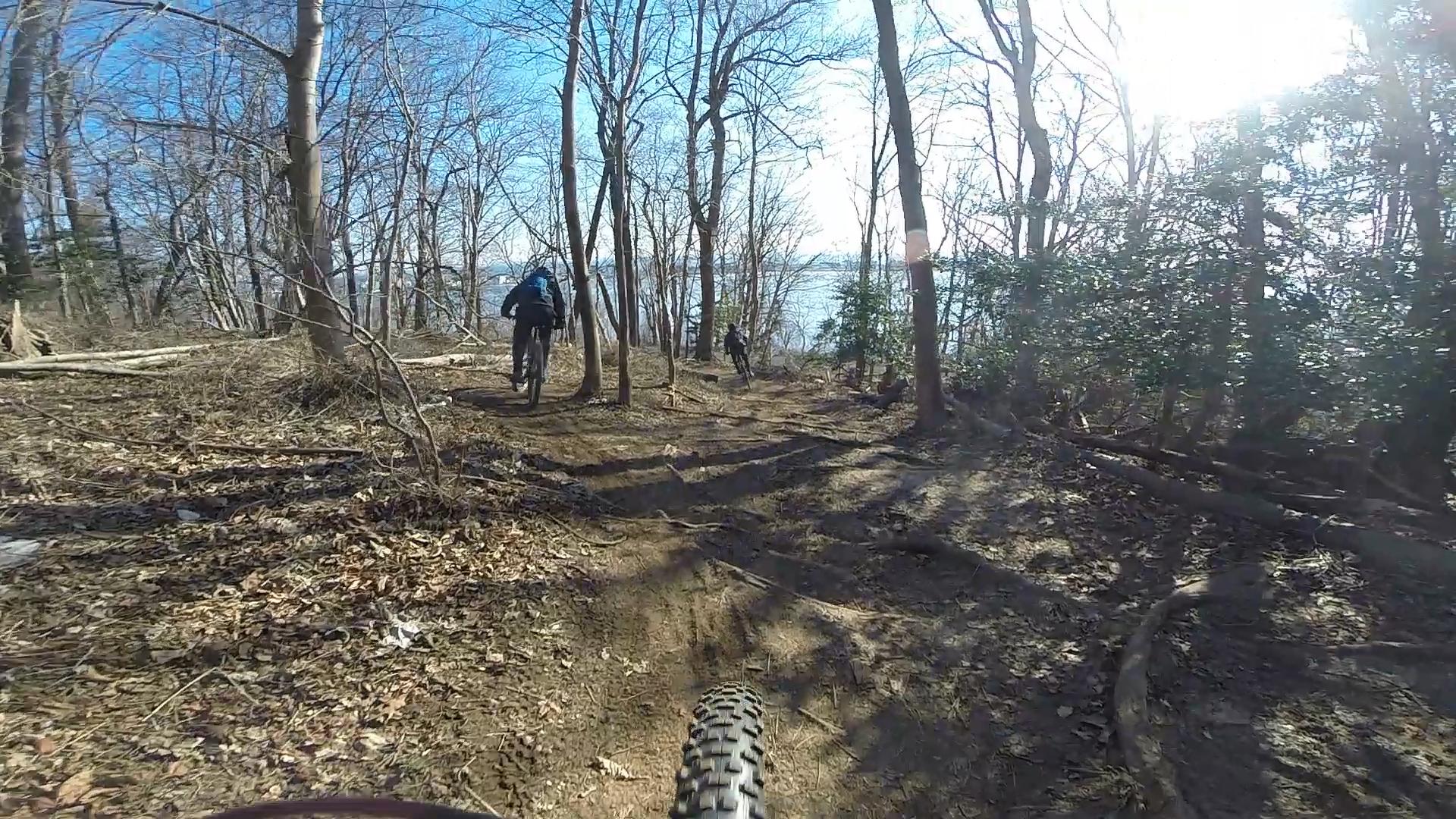 Two mountain bikers navigate a dirt trail surrounded by trees in a wooded area. The scene captures a bright, sunny day with clear blue skies, as the bikers ride downhill near a body of water in the background. The foreground shows part of a bike's tire and the rugged trail, covered with fallen leaves and scattered branches. Hartshorne Woods Park mountain bike trail.