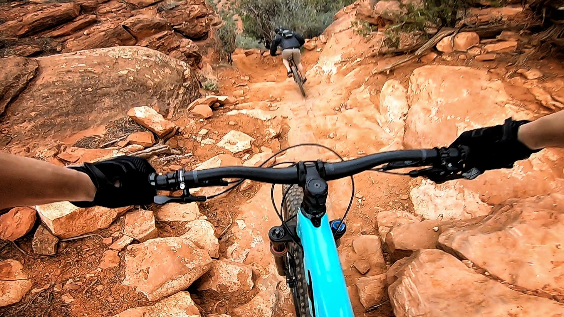 A first-person perspective view of a mountain biker navigating a rocky downhill trail. The handlebars of a blue mountain bike are in focus, with hands in gloves gripping them. In the background, another biker can be seen descending the rugged terrain. The landscape features red dirt and scattered rocks, surrounded by sparse vegetation. Hiline mountain bike trail.