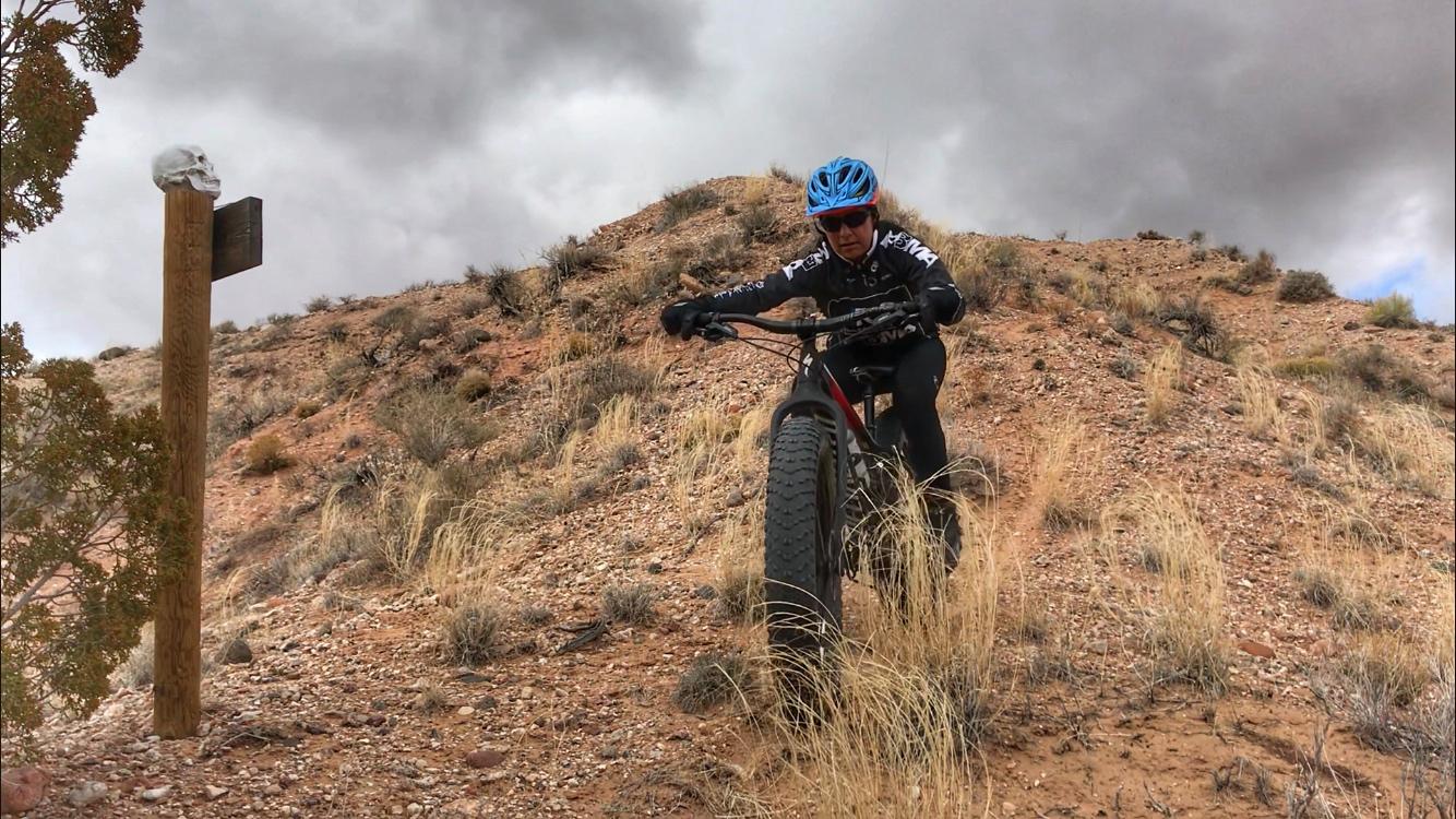 A cyclist wearing a blue helmet and black cycling gear climbs a sandy hill on a fat bike, surrounded by sparse vegetation and rocky terrain under a cloudy sky. A wooden signpost with a skull decoration is visible in the foreground. Mariposa Fat Bike Trails mountain bike trail.