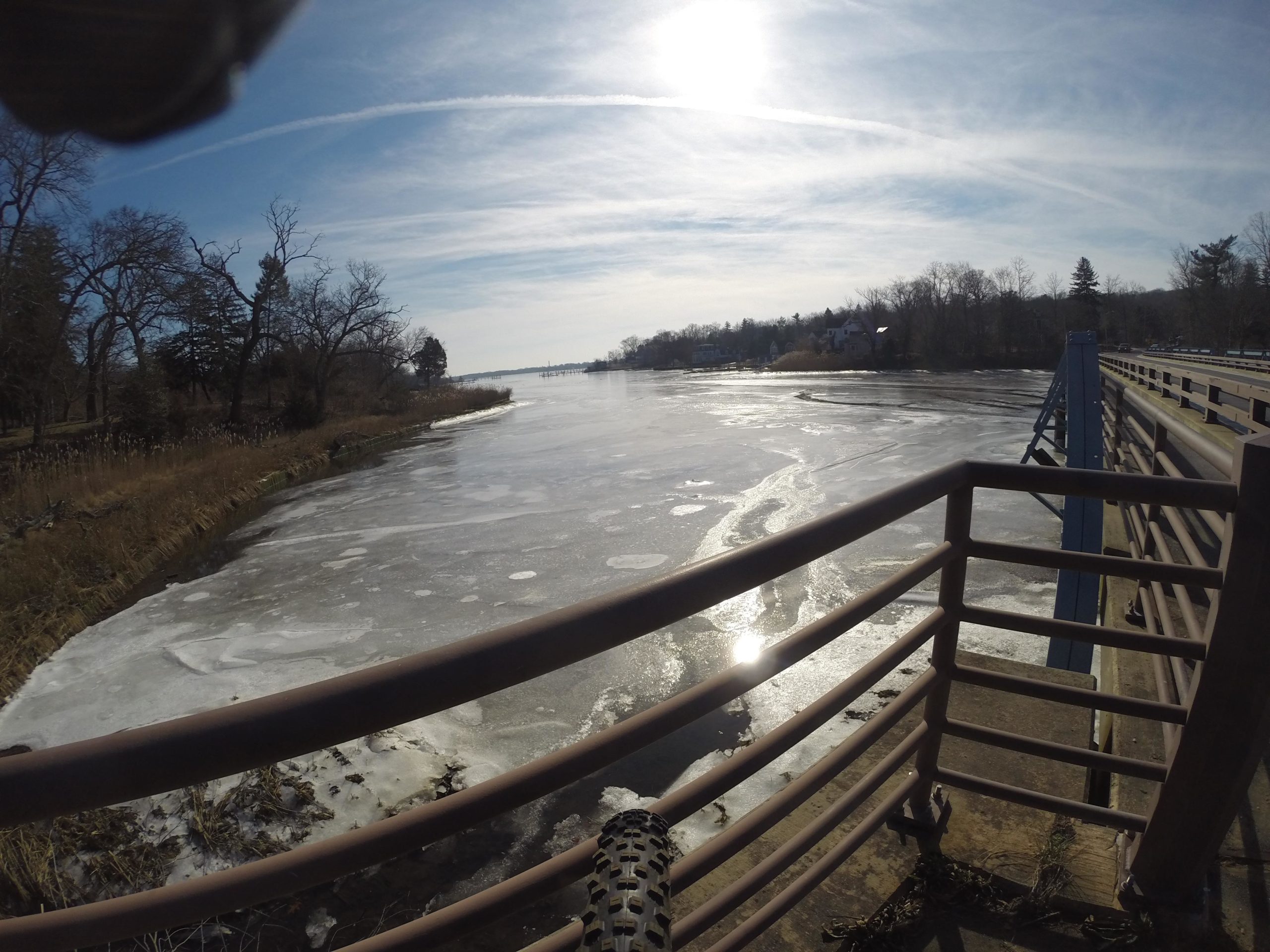 A scenic view of a frozen river taken from a bridge, with trees lining the banks and a bright sun shining in a clear blue sky. The water shows patches of ice and a few ripples, reflecting the sunlight. Huber Woods mountain bike trail.