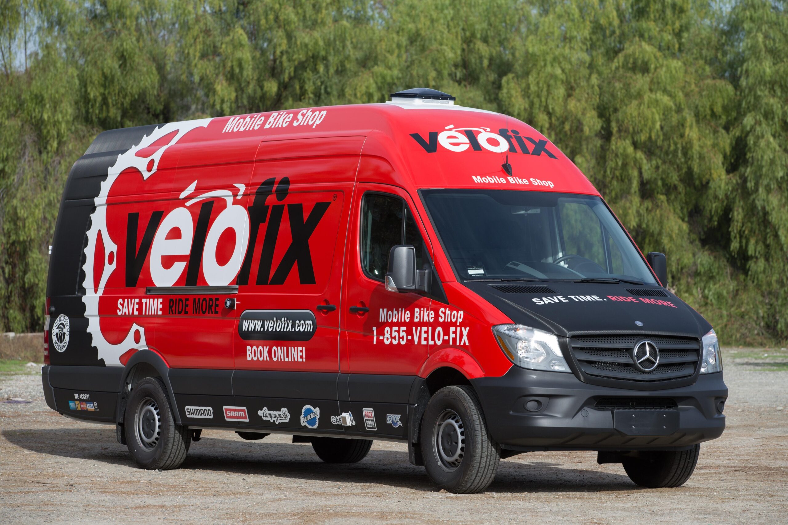 A bright red mobile bike shop van with the logo "vélofix" prominently displayed on the side. The van features text that reads "SAVE TIME. RIDE MORE." along with contact information and website details. Surrounding the van are green trees and a gravel surface, indicating an outdoor setting.