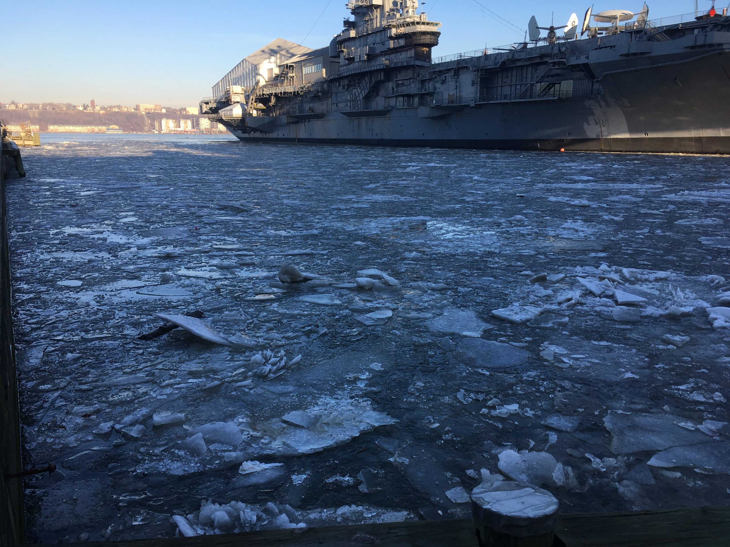 A frozen river with scattered ice floes, in the foreground, next to a large naval ship docked at the edge. In the background, a shoreline with buildings can be seen under a clear blue sky. West Street Greenway mountain bike trail.