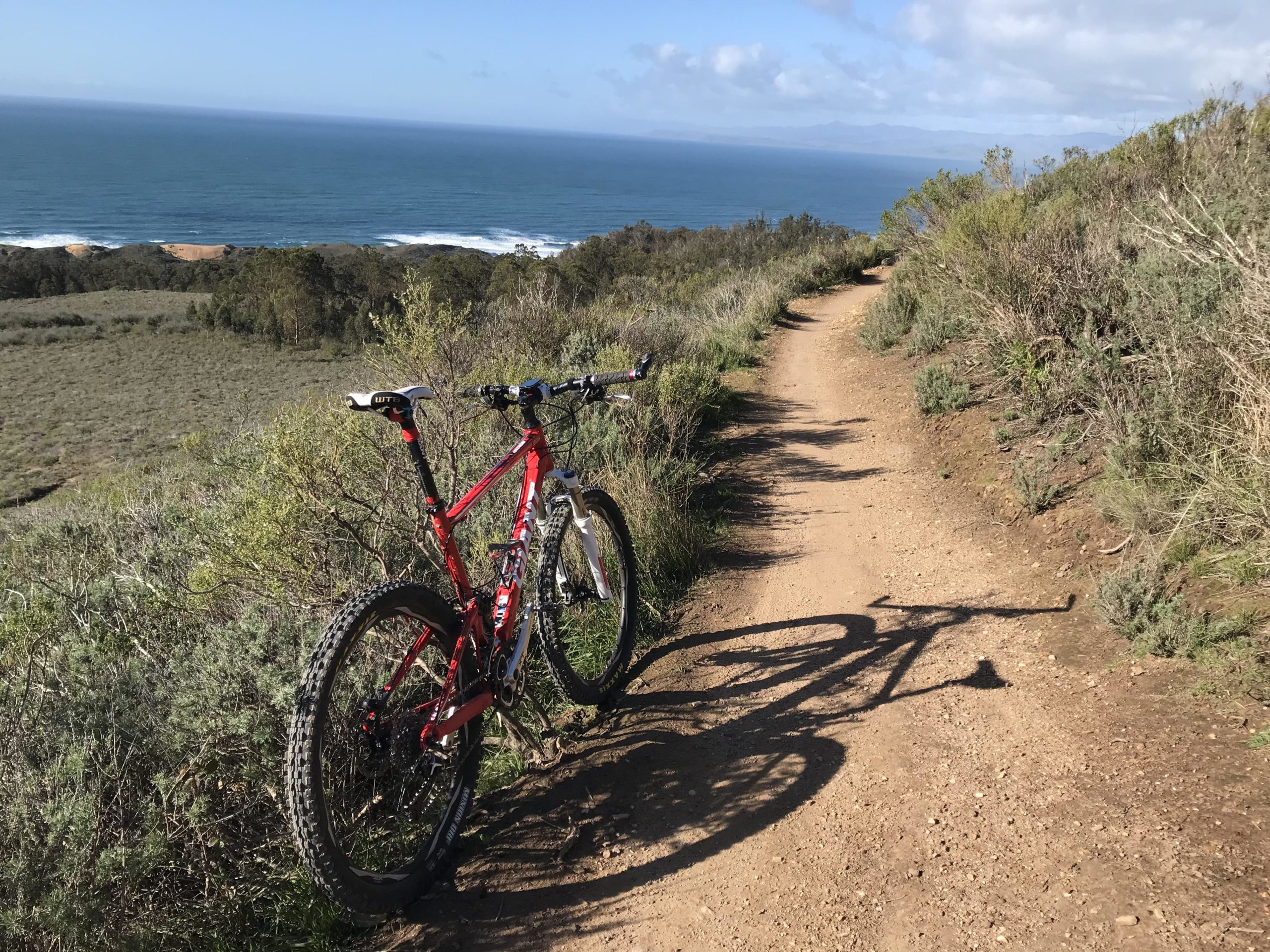 A red mountain bike leaning against a grassy hillside, with a dirt trail curving along the landscape. In the background, the view opens up to the ocean with waves crashing on the shore under a blue sky with scattered clouds. Montana De Oro mountain bike trail.
