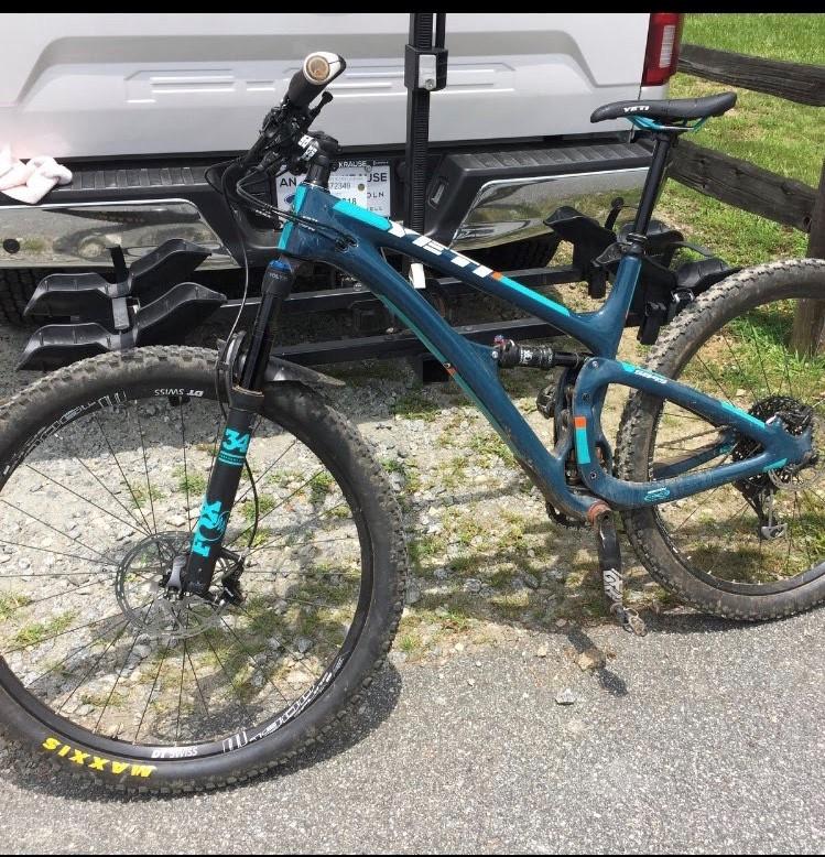 Yeti SB4.5c: A blue mountain bike is parked on gravel next to a pickup truck. The bike features large wheels with knobby tires, a front suspension fork, and a dropper seat post. The background includes grassy areas and a wooden fence.