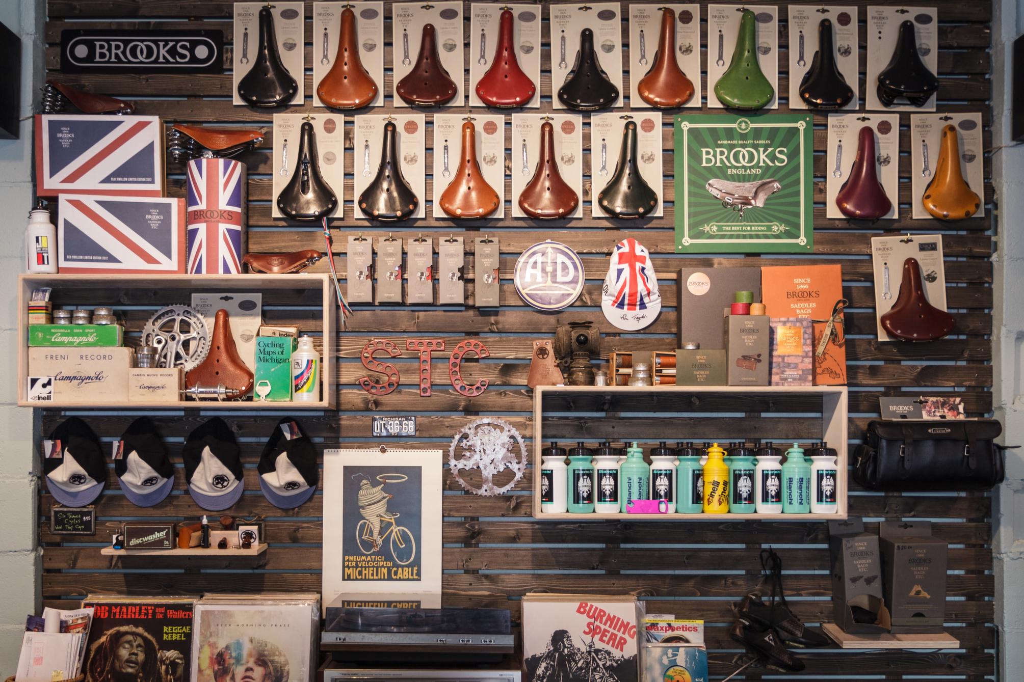 A vintage-style display wall featuring a variety of bicycle accessories, including multiple Brooks saddles, vintage packaging, caps, water bottles, and cycling memorabilia. The arrangement showcases items against a wooden backdrop, highlighting Brooks branding and a mix of colorful and metallic finishes.