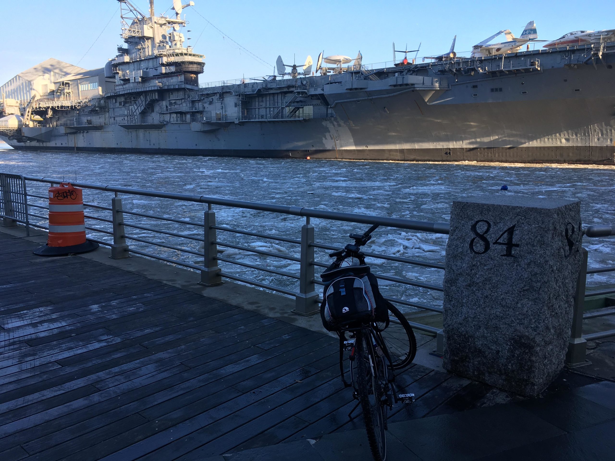 A large ship moored along a river with visible icy water, alongside a wooden walkway. In the foreground, a bicycle is parked next to a stone post marked with the number 84, and an orange traffic barrel is seen nearby. Bright blue sky adds contrast to the scene. West Street Greenway mountain bike trail.