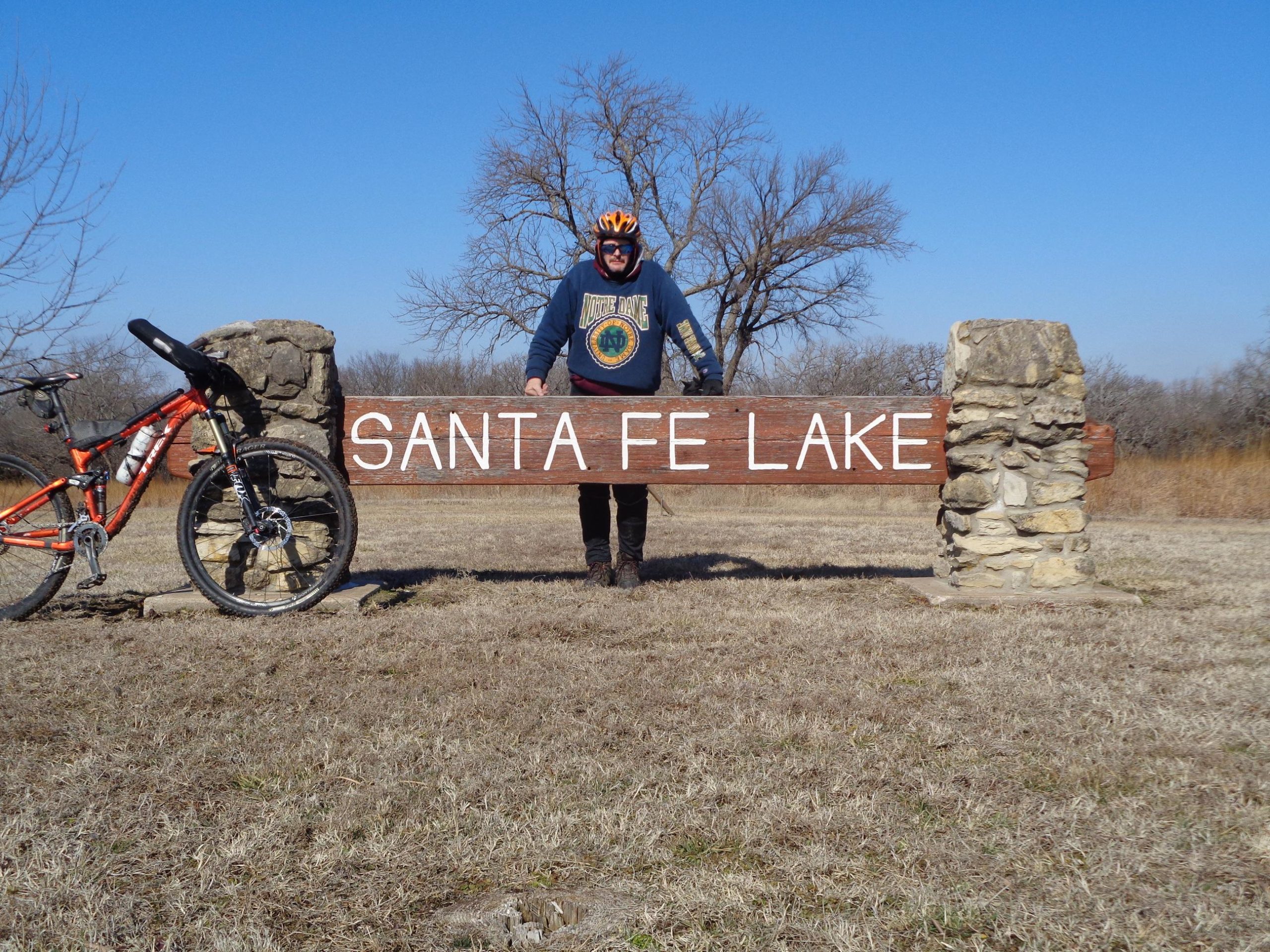 A person in a blue sweatshirt and helmet stands next to a wooden sign that reads "SANTA FE LAKE." An orange mountain bike leans against the sign. The background features a clear blue sky and sparse trees, with grassy terrain visible in the foreground. Santa Fe Lake Trail mountain bike trail.