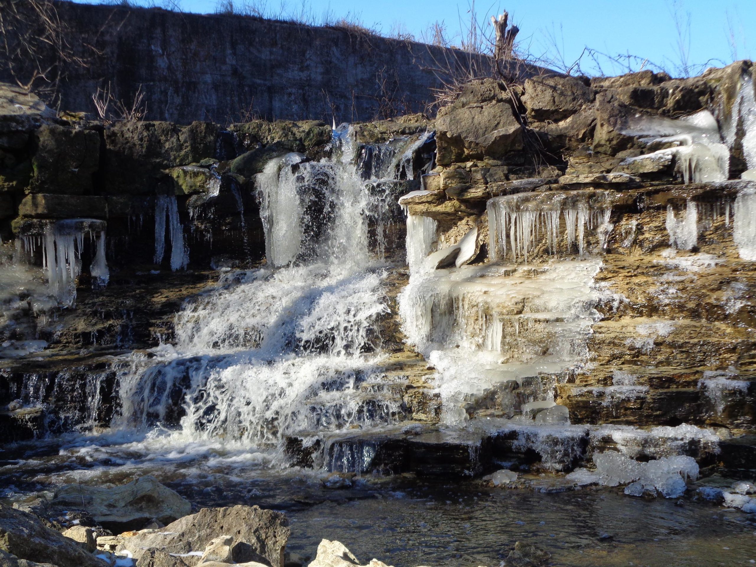 A frozen waterfall cascading over rocky terrain, with icicles hanging from the ledges and a clear blue sky in the background. Santa Fe Lake Trail mountain bike trail.