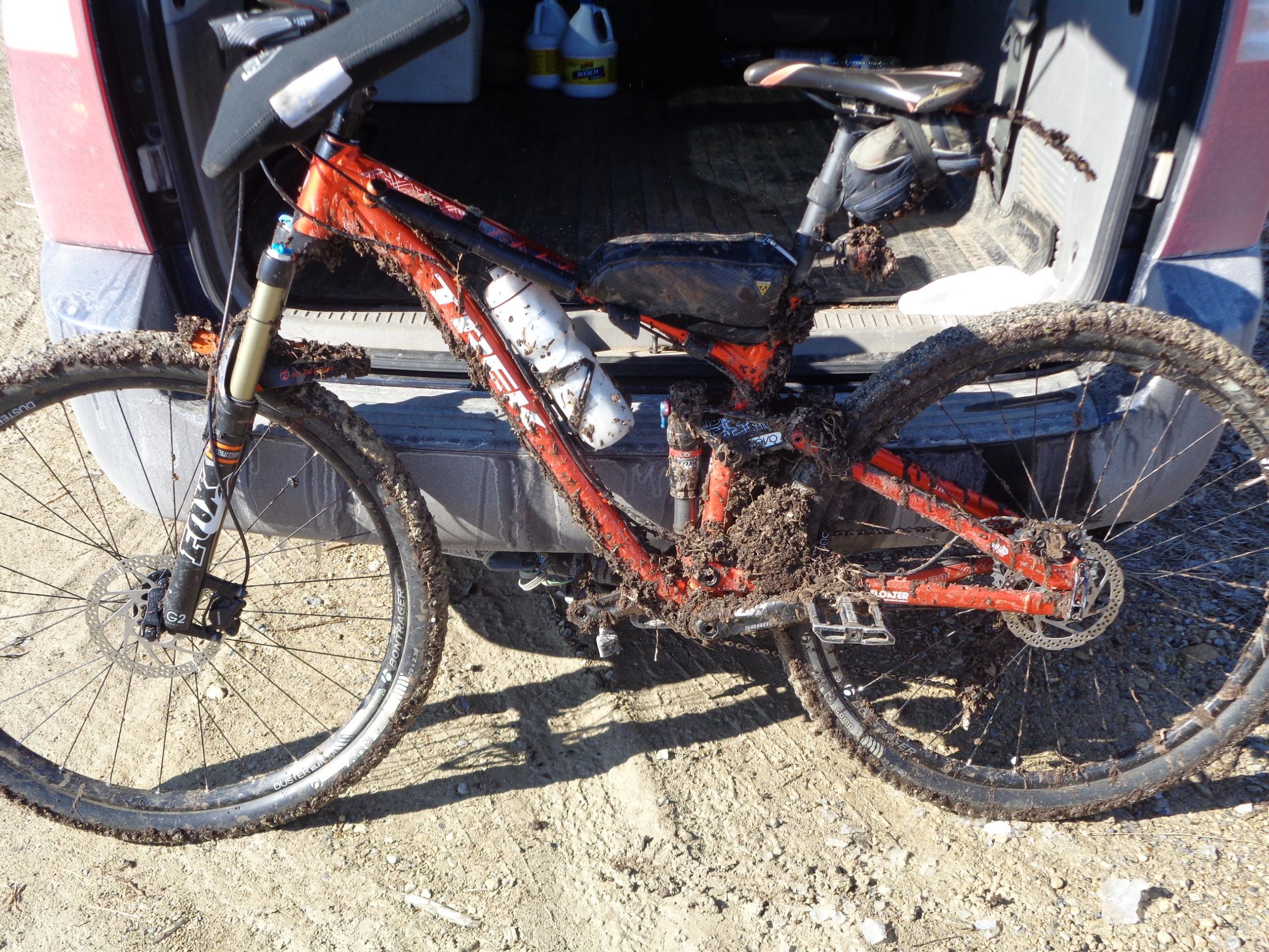 A muddy mountain bike leaning against the back of a vehicle, showcasing thick layers of mud on its frame, tires, and components. The bike has an orange color with various accessories attached, including a water bottle holder and a rear suspension. The background shows a sandy terrain and the interior of the vehicle. Santa Fe Lake Trail mountain bike trail.