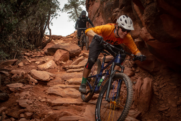 Two mountain bikers navigate a rocky trail in a rugged outdoor setting. One rider, wearing an orange shirt and a white helmet, is focused on the path ahead as he maneuvers through the uneven terrain. The second rider, dressed in black, follows closely behind. The landscape features red rock formations and sparse vegetation, typical of a desert environment.
