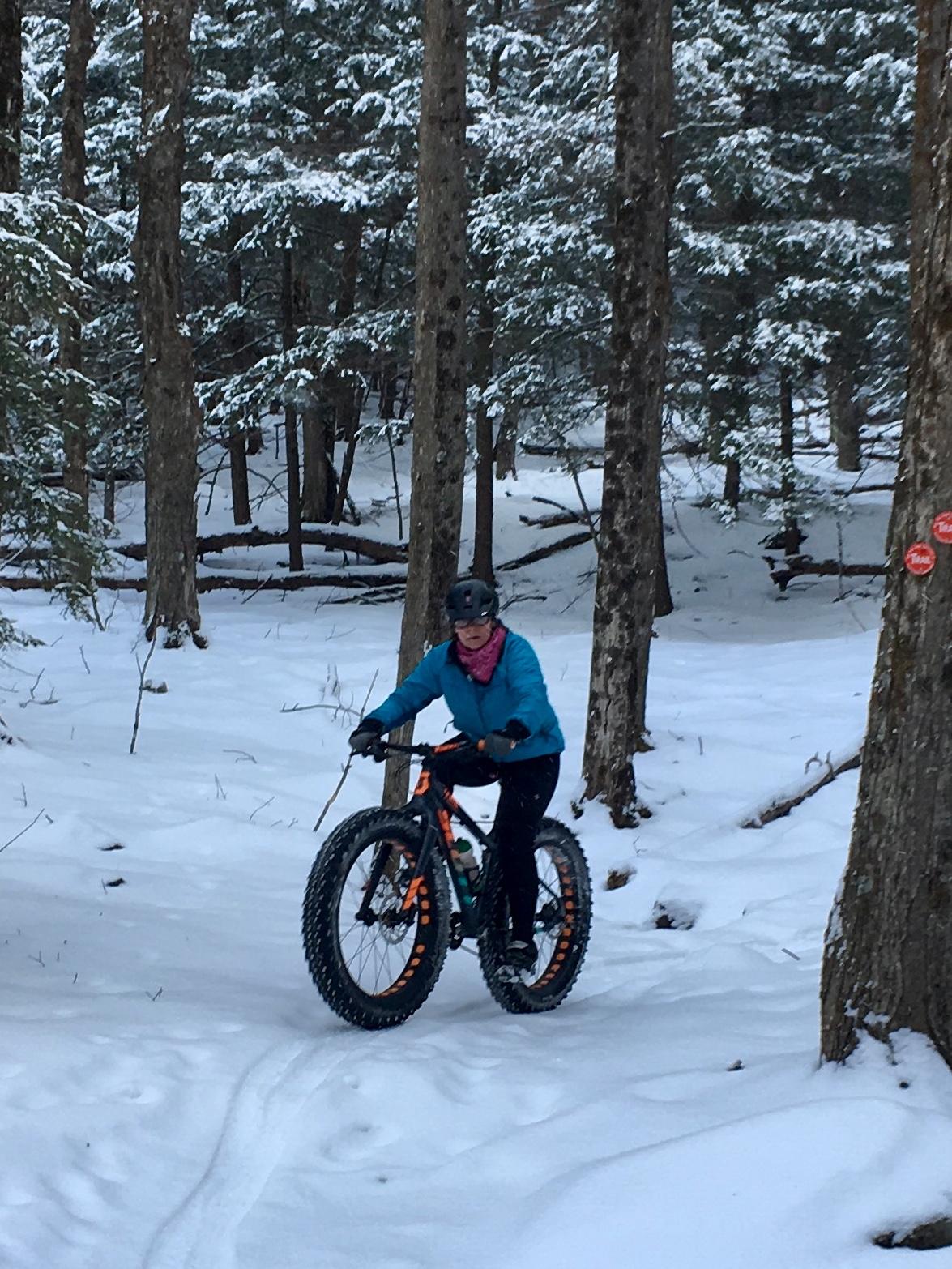 A person riding a fat bike on a snow-covered trail in a forest, surrounded by tall trees. The rider is wearing a blue jacket, a pink scarf, and a helmet. Snow is visible on the ground and the branches of the trees. Elm Ridge mountain bike trail.