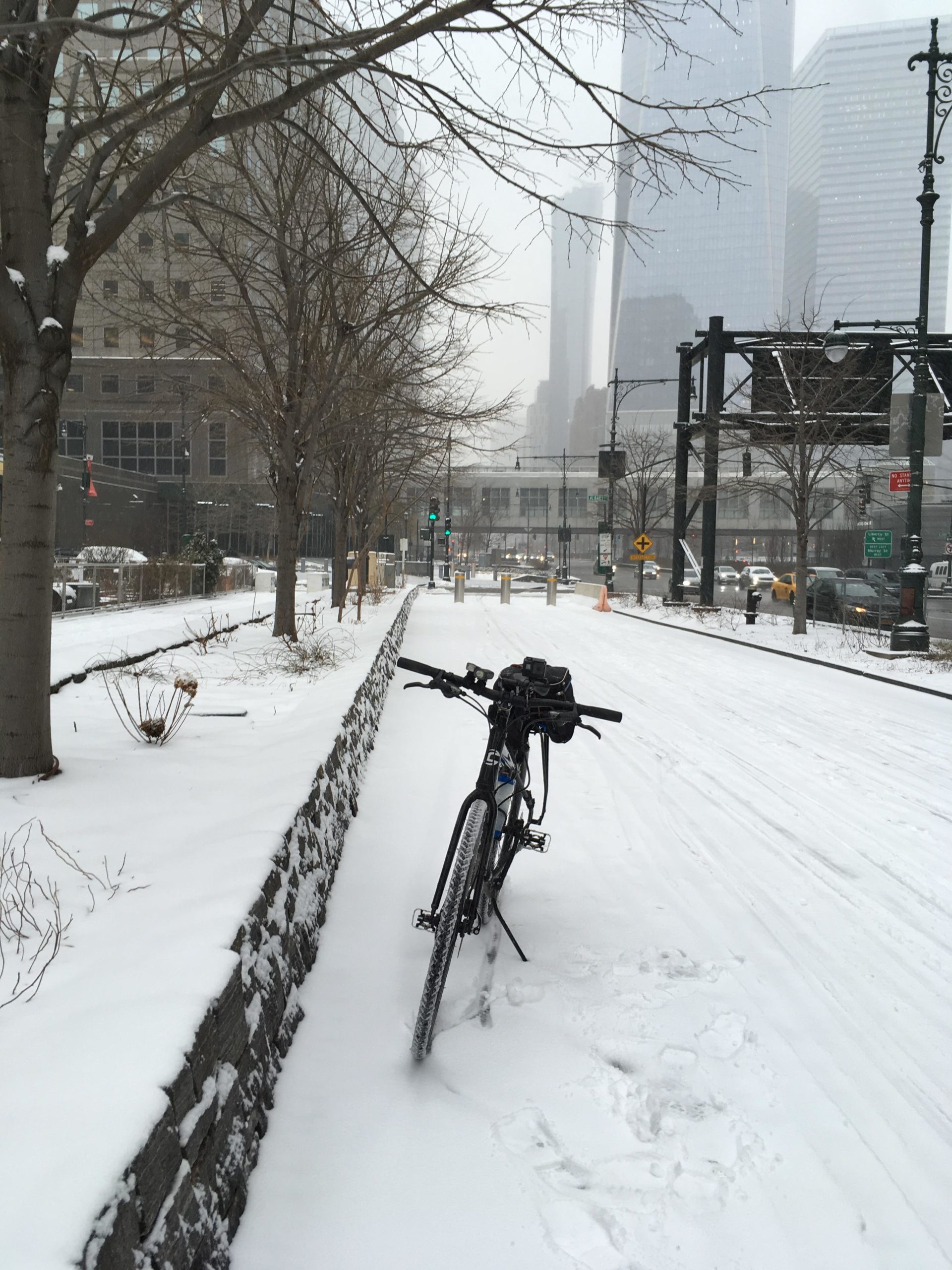 A bicycle rests against a stone wall on a snow-covered path, surrounded by barren trees. The scene is enveloped in a light snowfall, with tall buildings faintly visible in the background through a misty atmosphere. Green traffic lights and street signs can be seen along the edge of the path, indicating an urban setting in winter. West Street Greenway mountain bike trail.