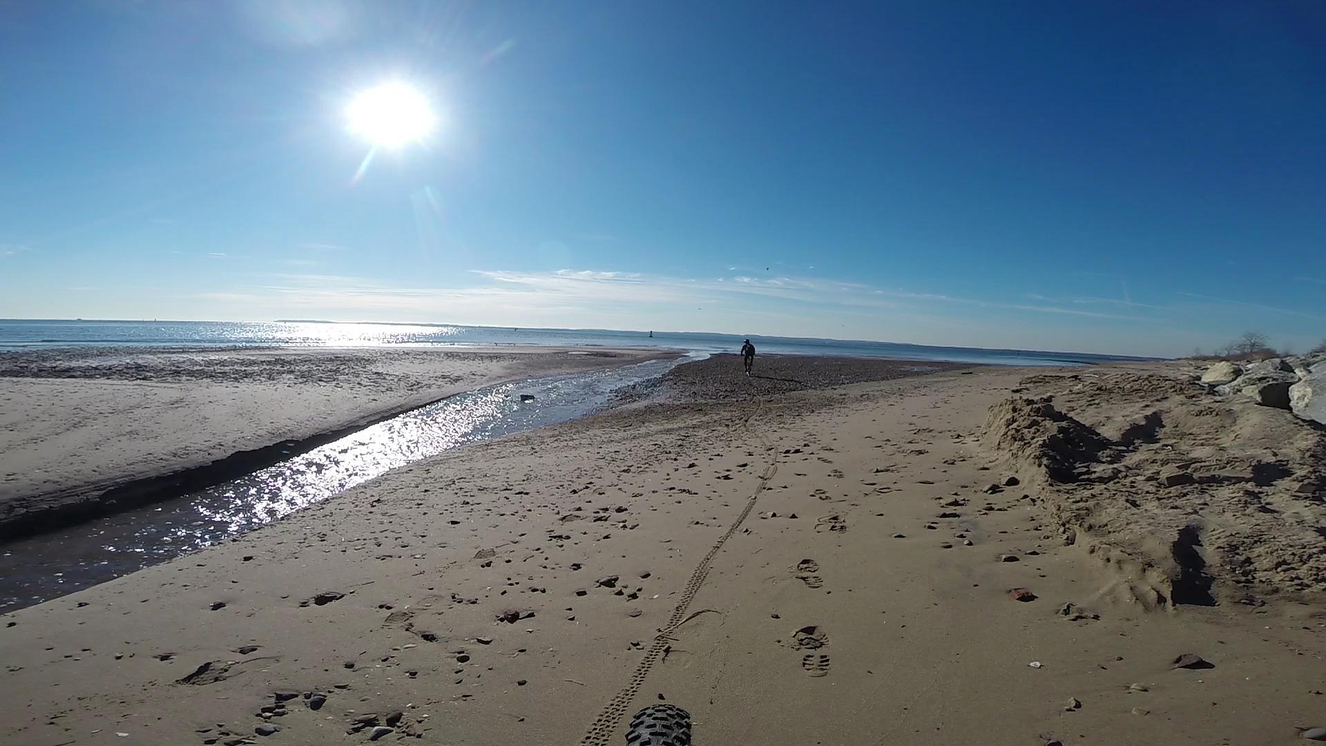 A scenic beach view on a sunny day, featuring a clear blue sky and bright sunlight reflecting on the ocean. A sandy shoreline with scattered footprints leads toward a distant figure walking along the water’s edge. A small stream flows through the sand, and rocky formations are visible on the right side of the image. Wolfes Pond park mountain bike trail.