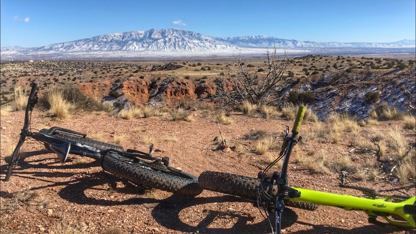 A mountain bike lying on a rocky landscape with sparse vegetation, overlooking a vast valley and snow-capped mountains in the background under a clear blue sky. Mariposa Fat Bike Trails mountain bike trail.