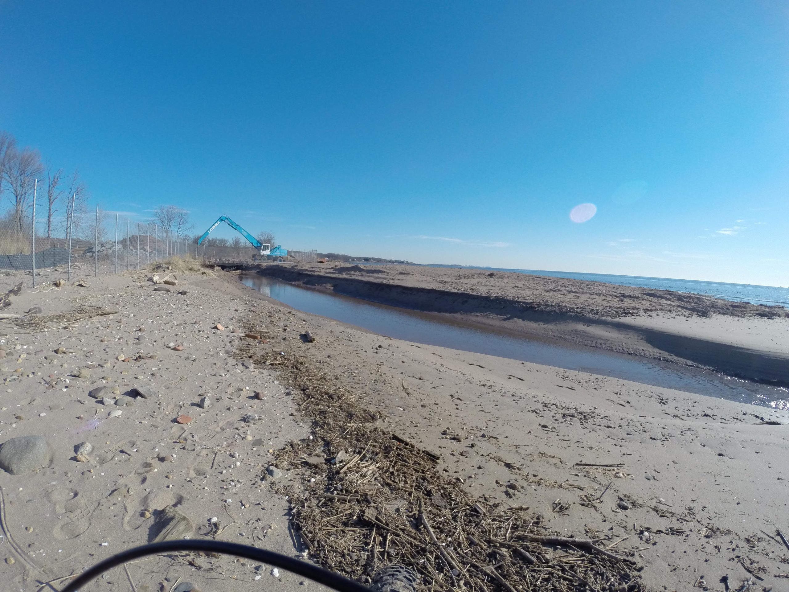 A sandy beach scene featuring a clear blue sky and calm water. In the background, there is a blue construction vehicle positioned near a fenced area, indicating ongoing work along the shoreline. The foreground includes scattered stones and debris along the water's edge. Wolfes Pond park mountain bike trail.
