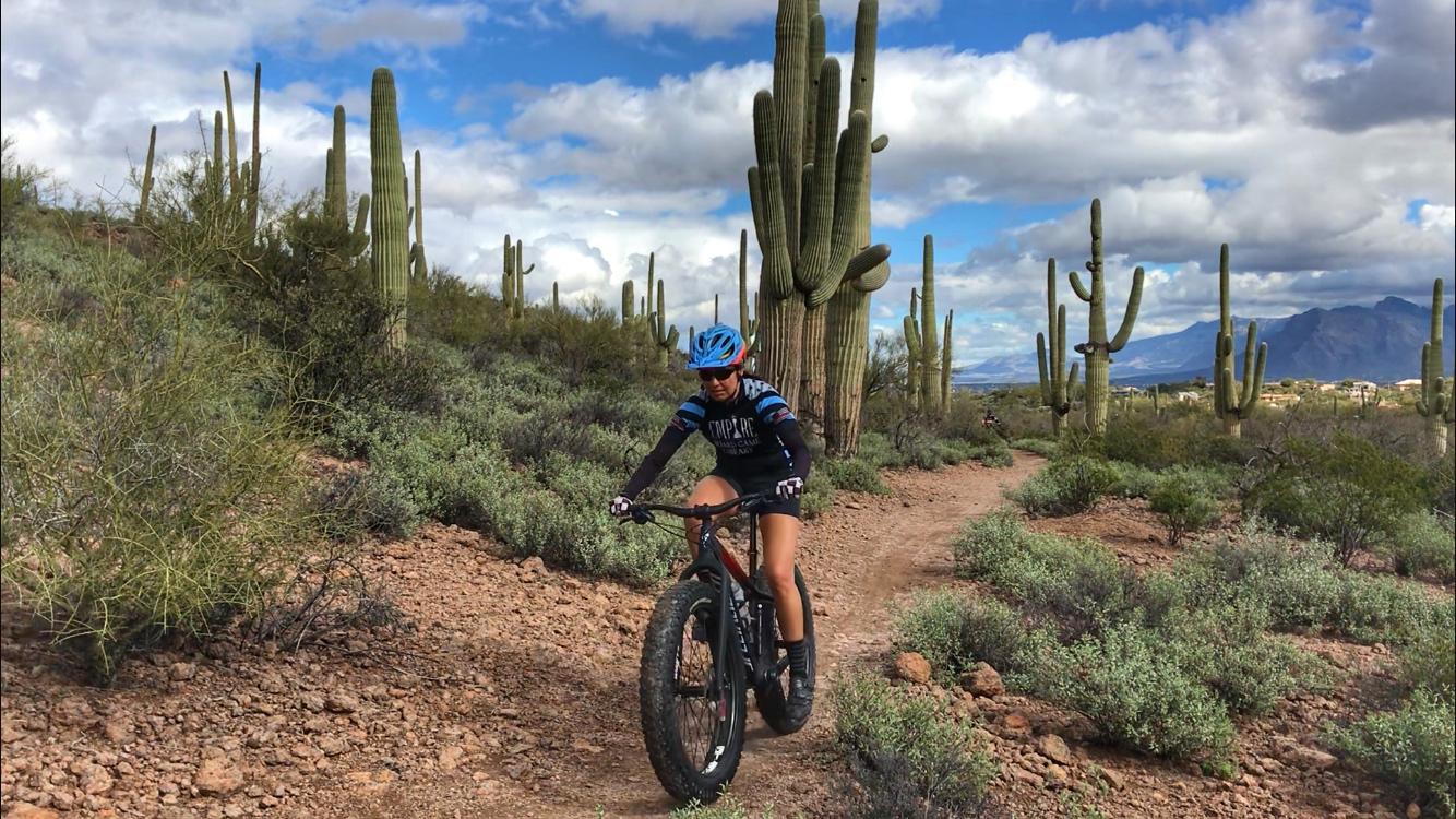 A person riding a fat bike on a dirt trail surrounded by cacti and shrubs in a desert landscape under a partly cloudy sky. Sweetwater Preserve mountain bike trail.