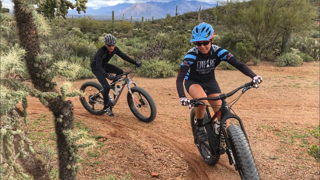 Two mountain bikers navigate a dirt trail in a desert landscape, surrounded by cacti and shrubs. One rider is wearing a blue helmet and a black and blue jersey, while the other is dressed in black attire. The background features rolling hills and mountains under a cloudy sky. Sweetwater Preserve mountain bike trail.