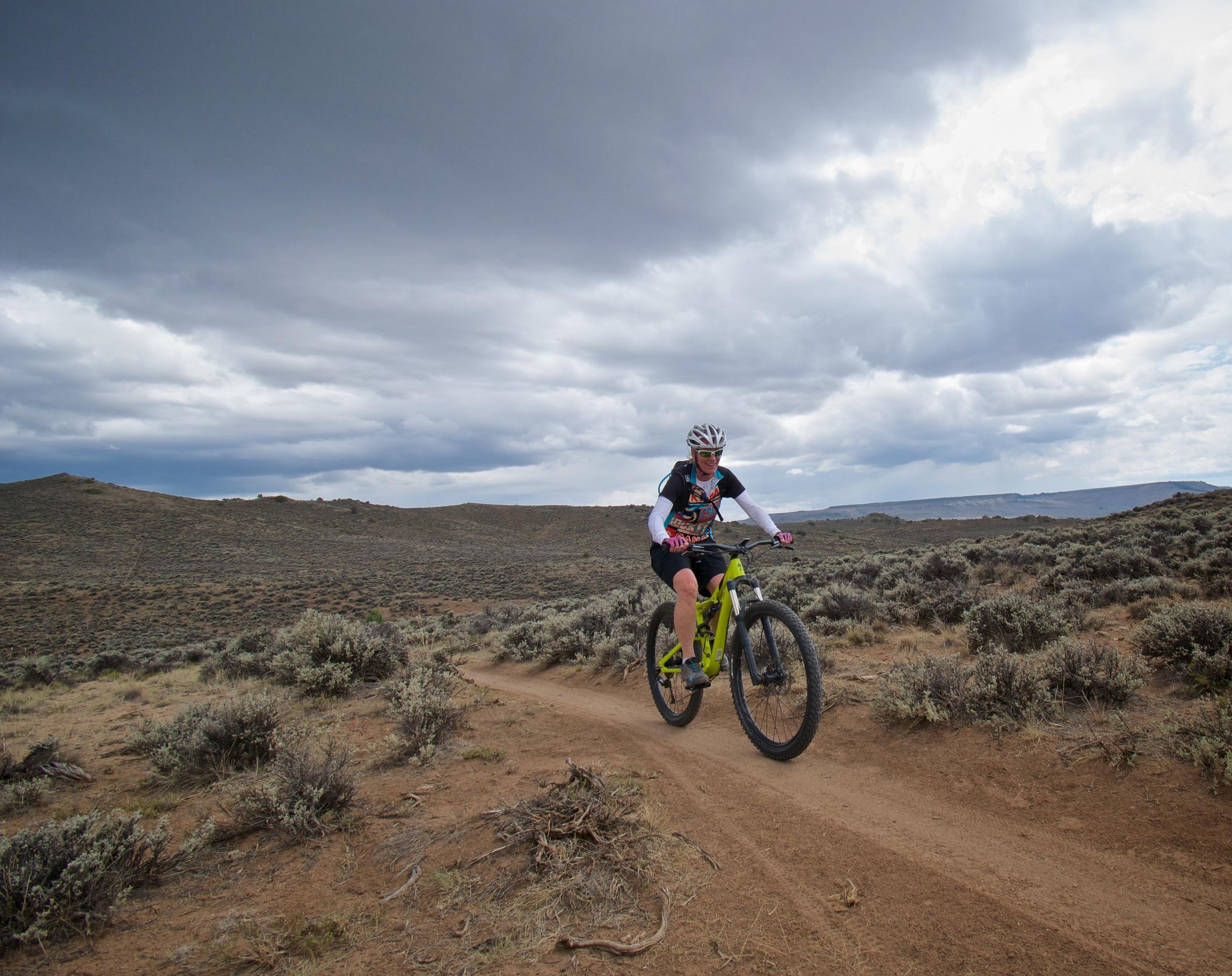 A mountain biker in a black and white outfit rides on a dirt trail through a rugged, bush-covered landscape under a cloudy sky. Hartman Rocks mountain bike trail.