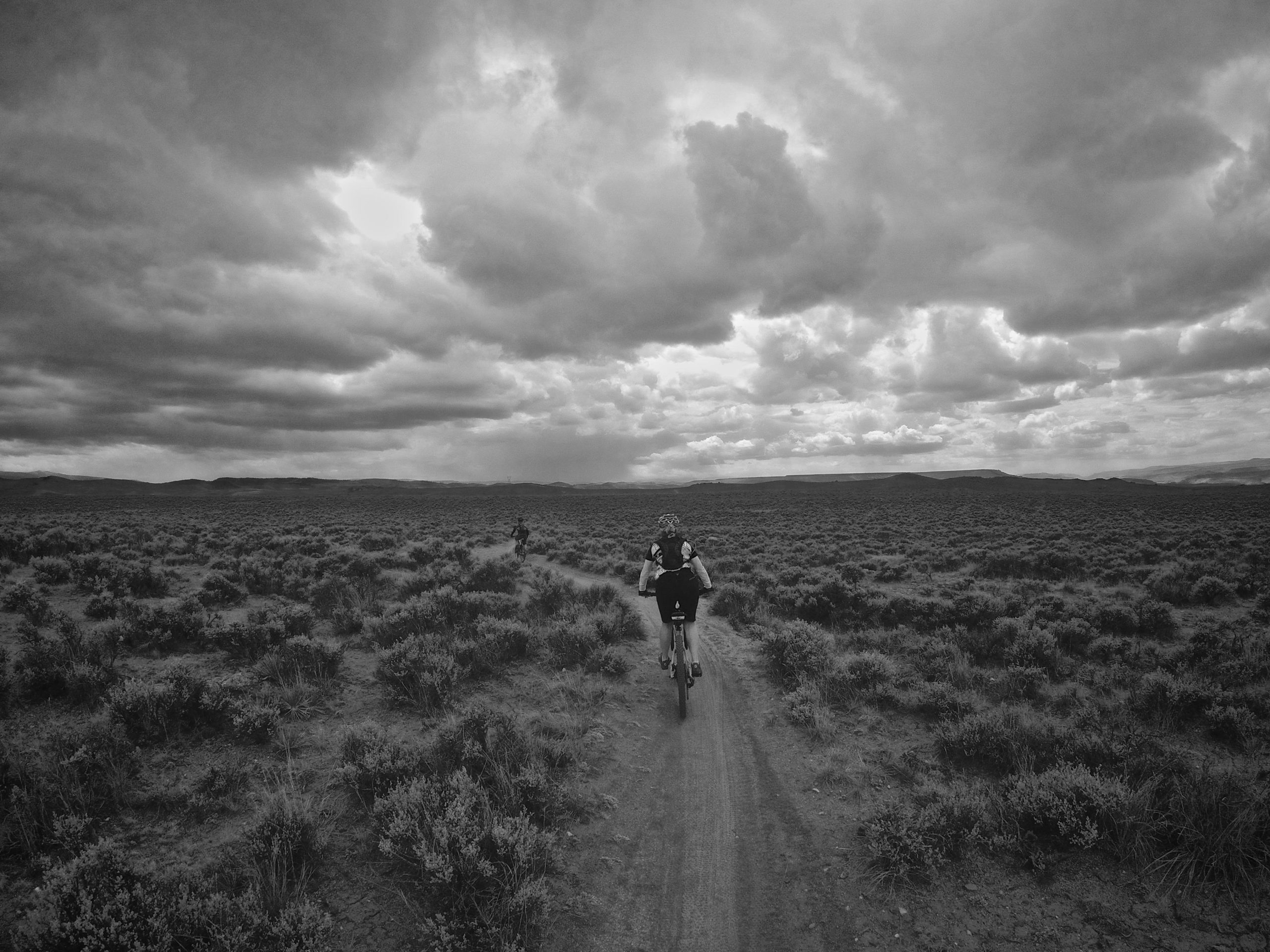 A monochrome image of two mountain bikers riding along a dirt trail in a vast, open landscape. The scene features a cloudy sky with dramatic cloud formations and a foreground of scrubby vegetation. The bikers appear small against the expansive background, which includes distant hills and a sense of remote wilderness. Hartman Rocks mountain bike trail.