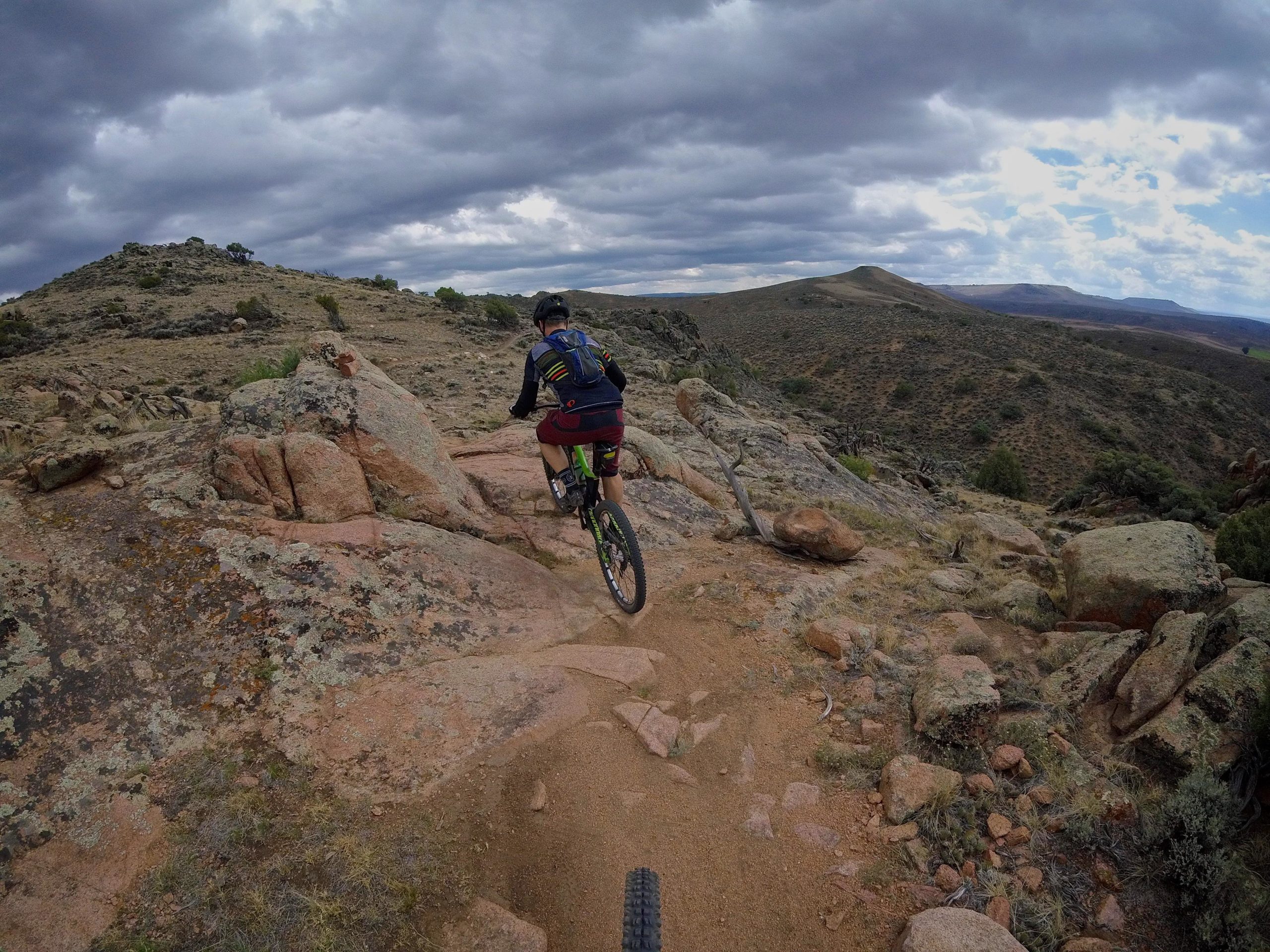 A mountain biker navigates a rocky trail through a rugged landscape, surrounded by hills and a cloudy sky. The terrain features patches of grass and scattered rocks, showcasing the challenges of off-road cycling. Hartman Rocks mountain bike trail.