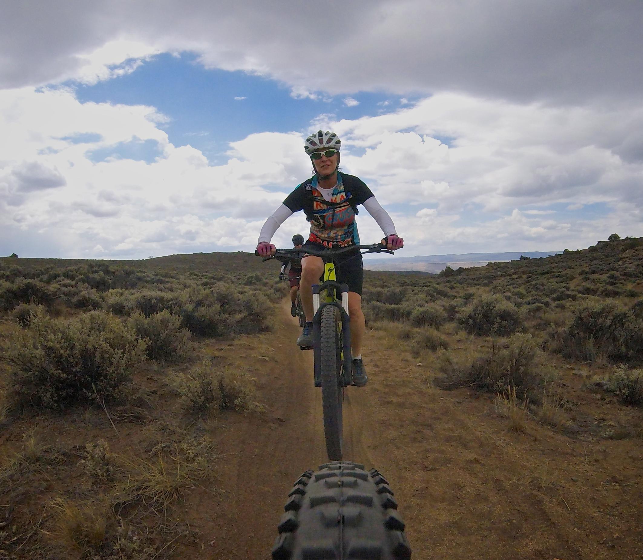 A mountain biker riding on a dirt trail surrounded by shrubs and hills, with a cloudy sky in the background. The cyclist is wearing a helmet and colorful jersey, and another biker is visible in the background. Hartman Rocks mountain bike trail.