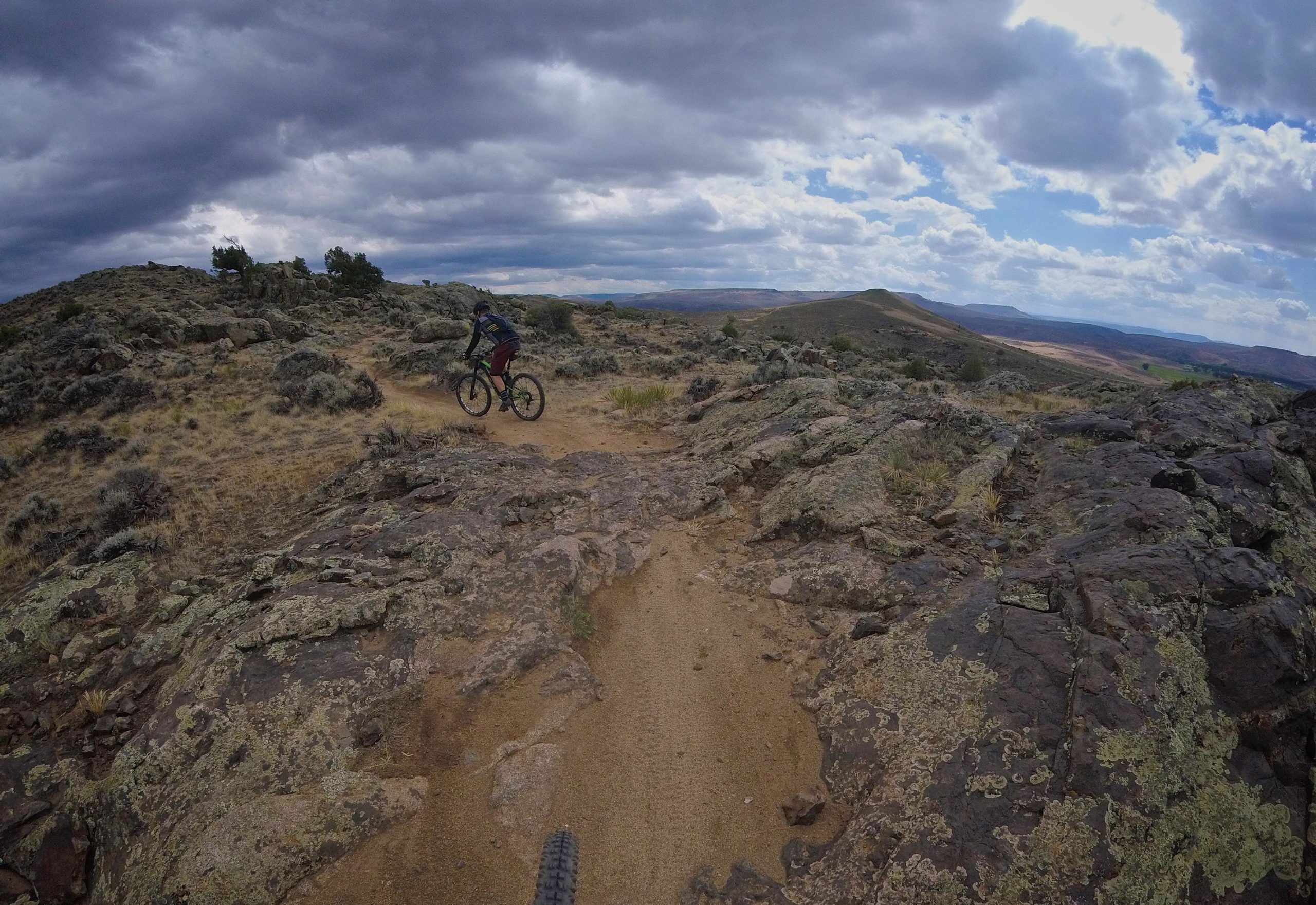 A person riding a mountain bike on a rocky trail, surrounded by rugged terrain and sparse vegetation under a cloudy sky. The landscape features hills in the background, creating a scenic view of an outdoor adventure environment. Hartman Rocks mountain bike trail.