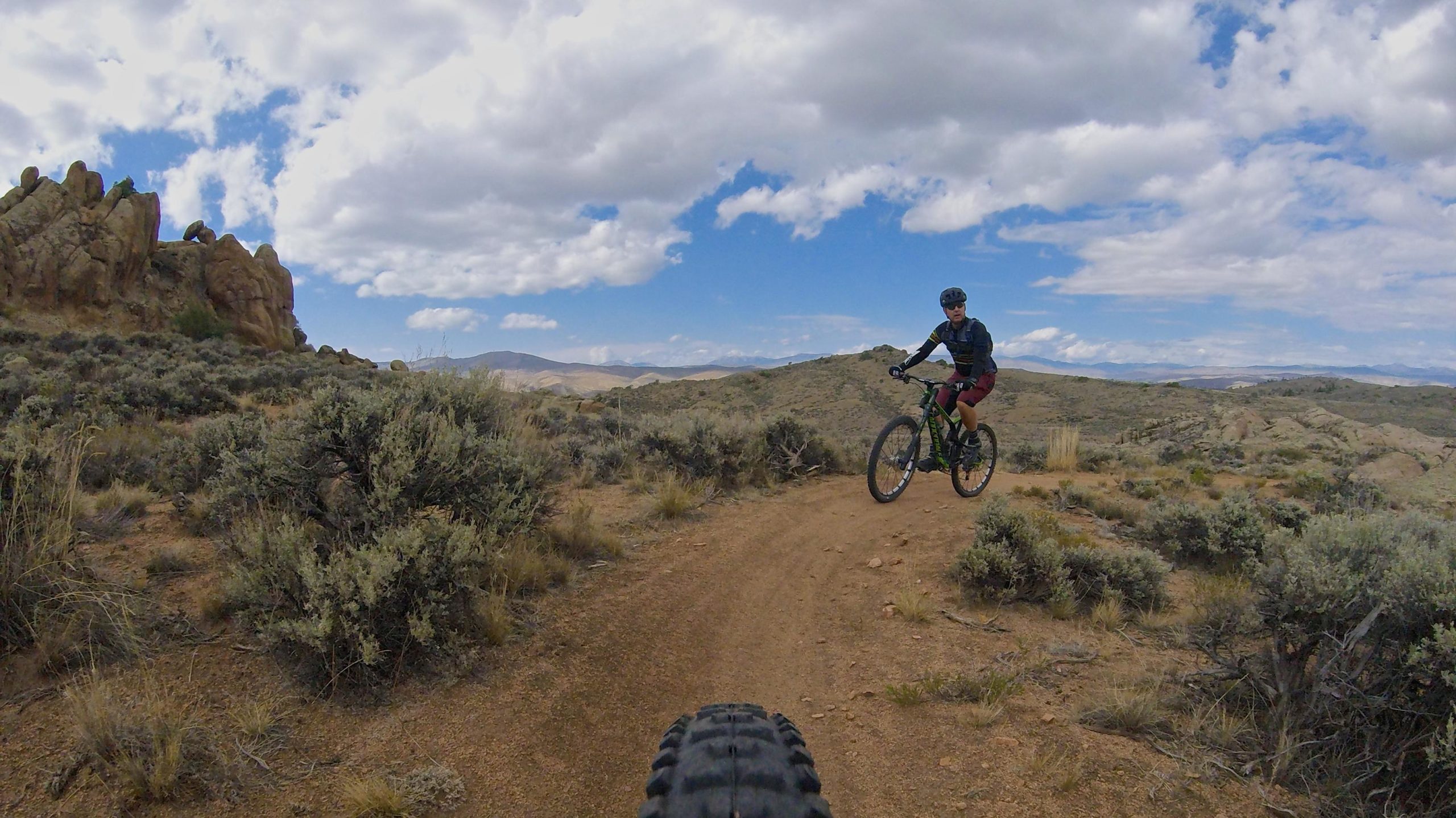 A mountain biker riding on a dirt trail in a rugged, outdoor landscape. The scene features rocky formations in the background and a mix of shrubs and vegetation along the path. The sky is partly cloudy, with patches of blue visible. The image is taken from a low angle, emphasizing the bicycle tire in the foreground. Hartman Rocks mountain bike trail.