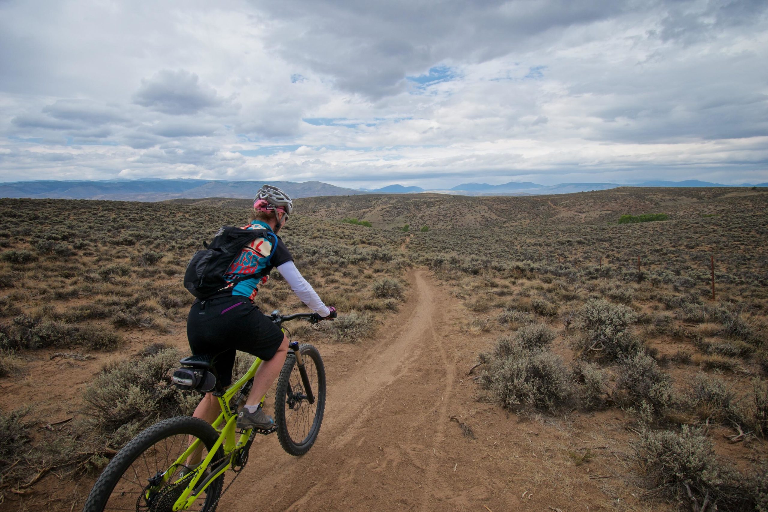 A person riding a mountain bike along a sandy trail in a rugged, open landscape with sparse vegetation and distant mountains under a cloudy sky. Hartman Rocks mountain bike trail.