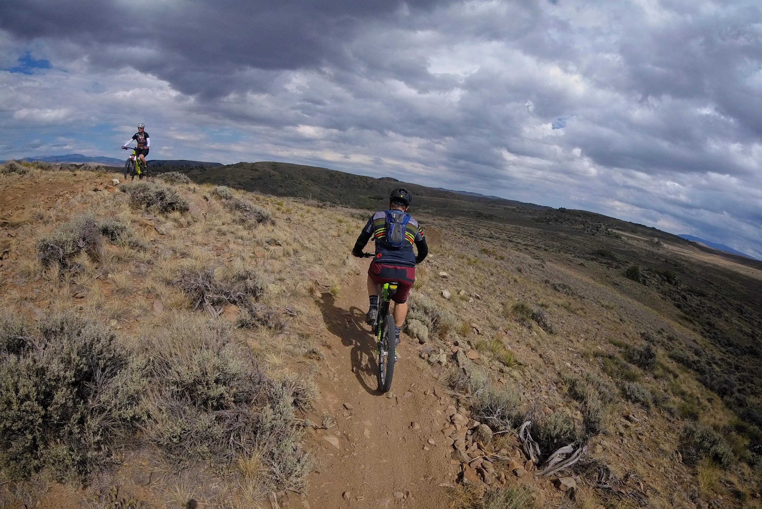 A mountain biker is riding along a dirt trail in a rugged landscape, surrounded by sparse vegetation and rolling hills under a cloudy sky. Another biker can be seen in the distance, also navigating the trail. Hartman Rocks mountain bike trail.