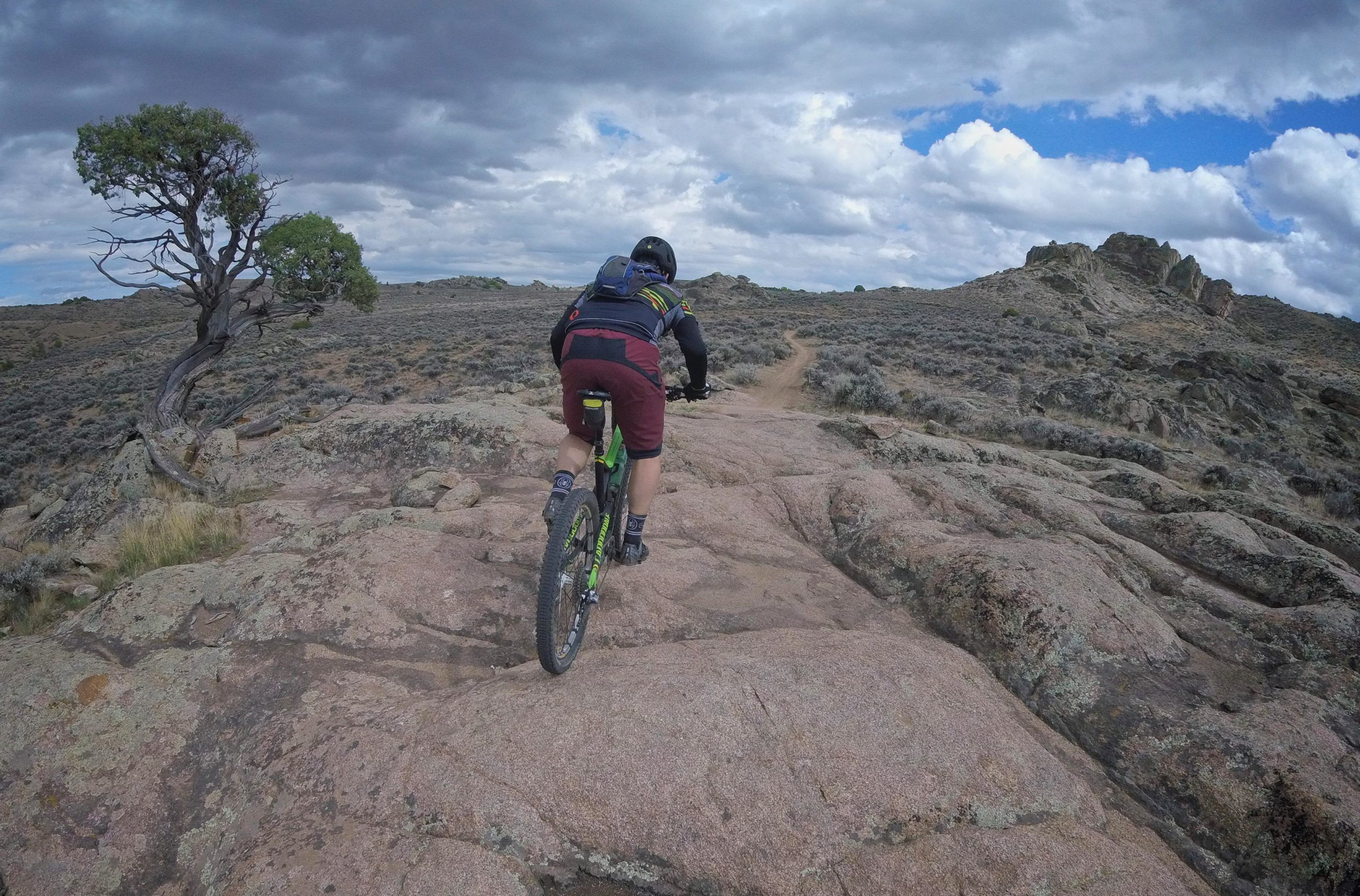 A mountain biker navigates a rocky trail in a rugged outdoor landscape, surrounded by sparse vegetation and a dramatic sky filled with clouds. A solitary tree stands to the side, adding to the natural scenery. Hartman Rocks mountain bike trail.