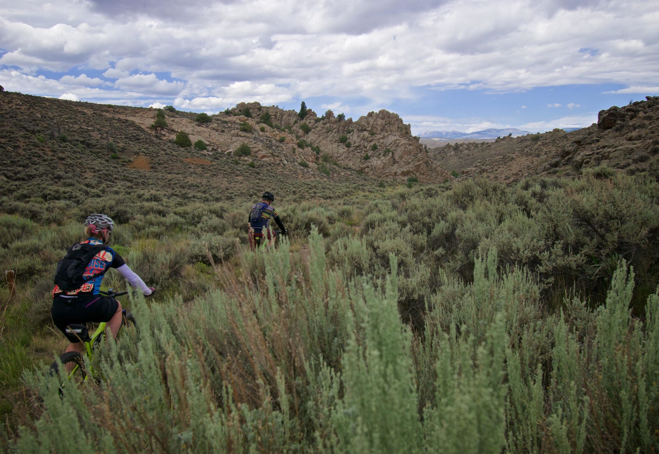 Two mountain bikers navigating a rocky, sagebrush-covered terrain under a partly cloudy sky. The landscape features rolling hills and rocky outcrops in the background, showcasing the beauty of outdoor cycling. Hartman Rocks mountain bike trail.