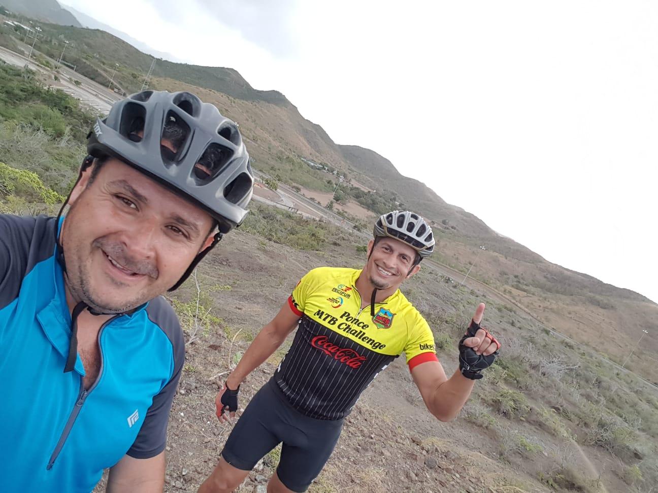 Two cyclists pose for a selfie on a rocky hillside with mountains in the background. One cyclist wears a blue shirt and a helmet, while the other is dressed in a yellow and black jersey and gives a thumbs-up gesture. The scene is captured under a cloudy sky. Los Pinchos mountain bike trail.