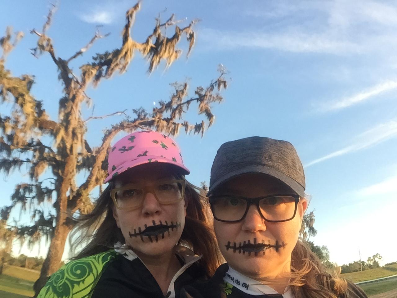 Two women pose for a photo outdoors, each with face paint resembling stitched lips. One woman wears a pink cap with a playful design, while the other sports a dark cap. In the background, a bare tree draped with Spanish moss is visible against a clear sky. The setting appears to be during the early evening with warm lighting. Bonnet Carre Spillway Trail mountain bike trail.
