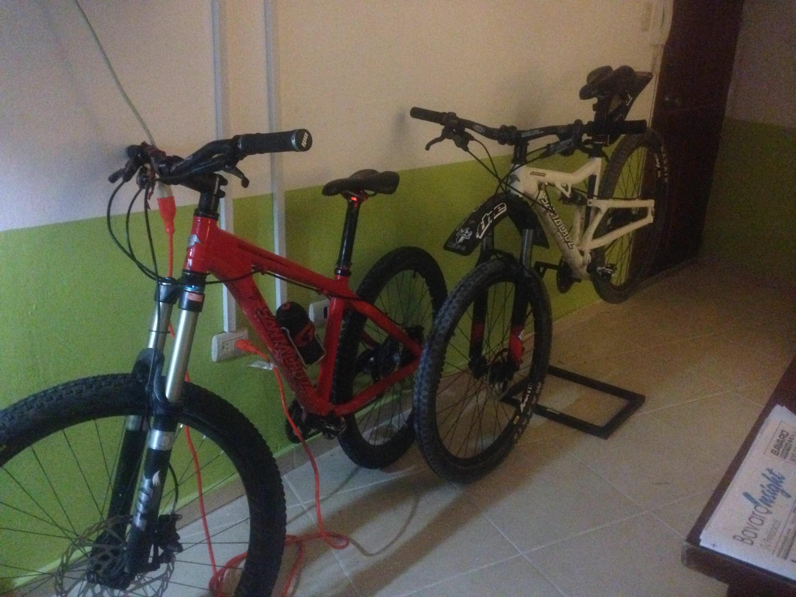 Santa Cruz Superlight: Two mountain bikes are leaning against a wall in a room. The bike on the left is red with a black and silver front fork, while the bike on the right is white with black accents. There are charging cables attached to the red bike, and the floor features light-colored tiles. The wall has a green lower section, and a wooden table is partially visible on the right.
