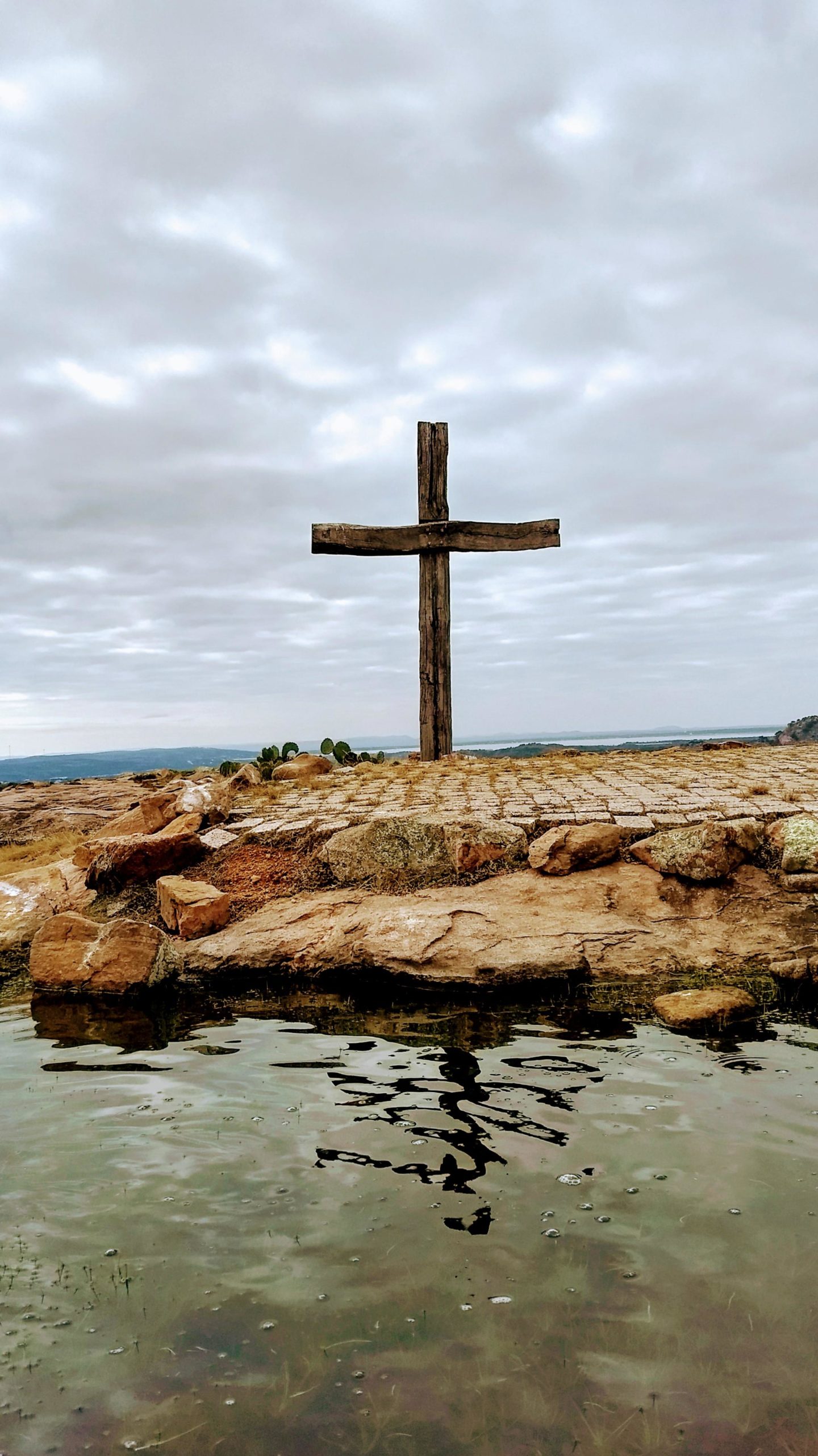 A wooden cross stands on rocky terrain overlooking a still body of water, with cloudy skies in the background. The cross is prominently placed on a flat stone surface, surrounded by natural elements. Reveille Peak Ranch mountain bike trail.